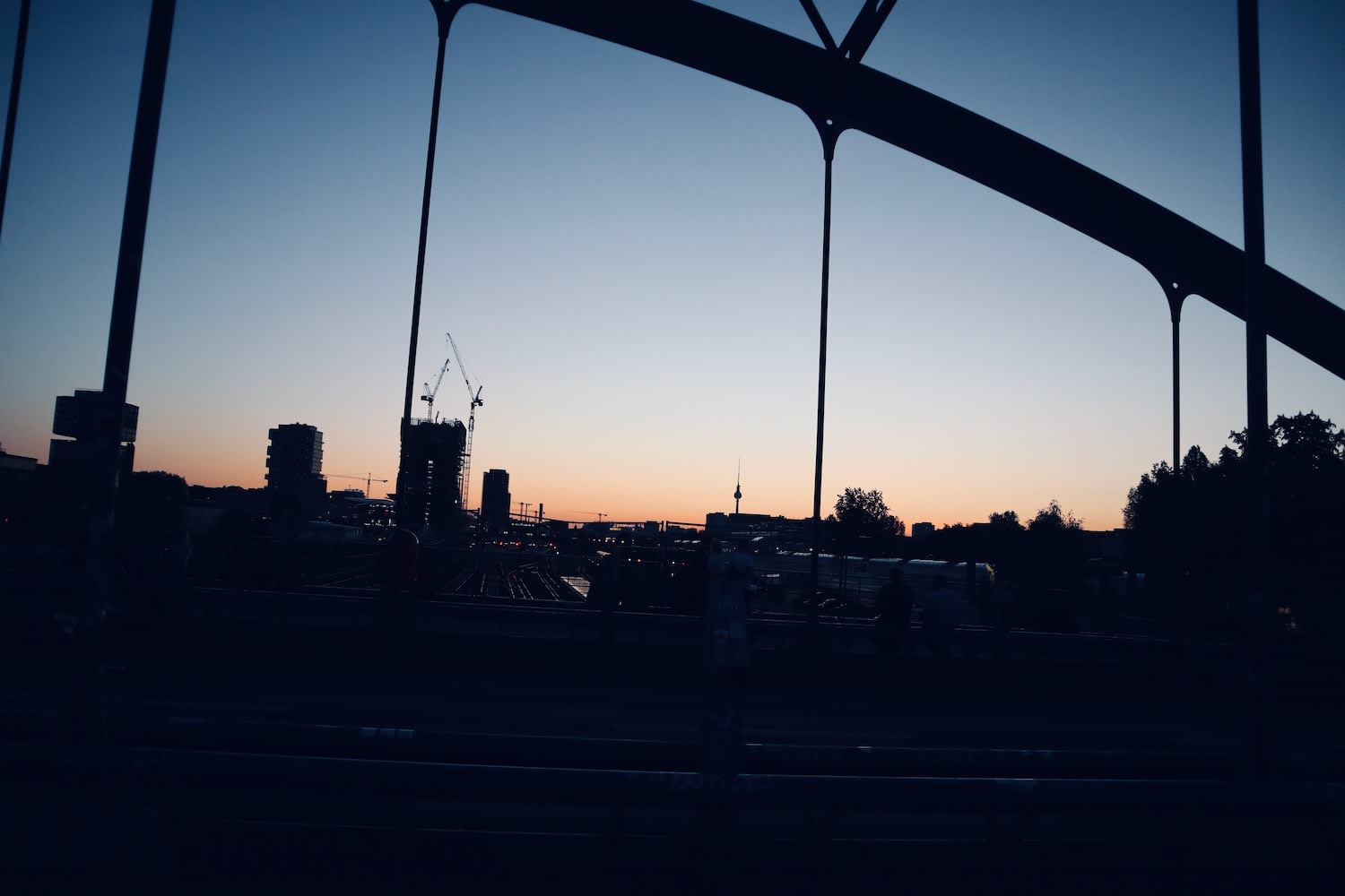 Twilight view from a bridge in Berlin, featuring urban outlines and construction cranes against a sunset backdrop, capturing life in Berlin.