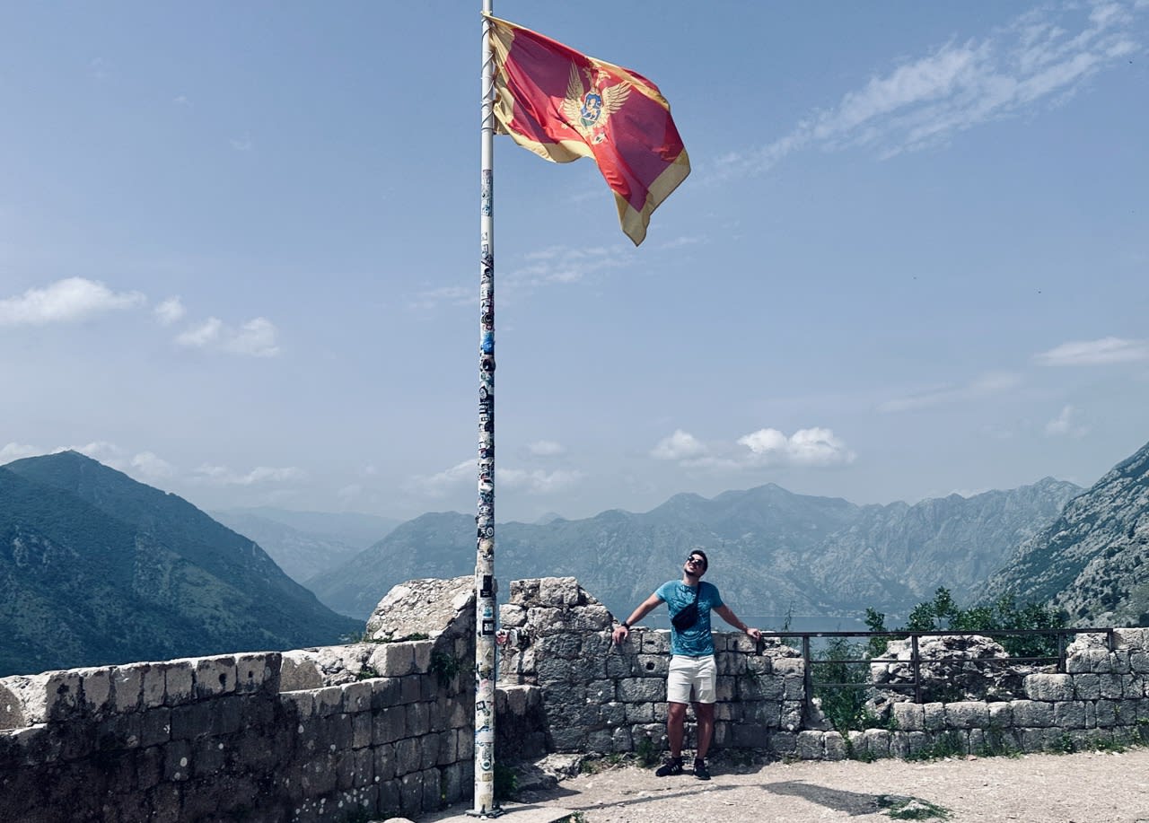Man standing next to a flagpole with the Montenegrin flag at a fortress in Kotor, Montenegro, enjoying a scenic view of the mountains, highlighting a travel destination accessible with a rent a car from Podgorica.
