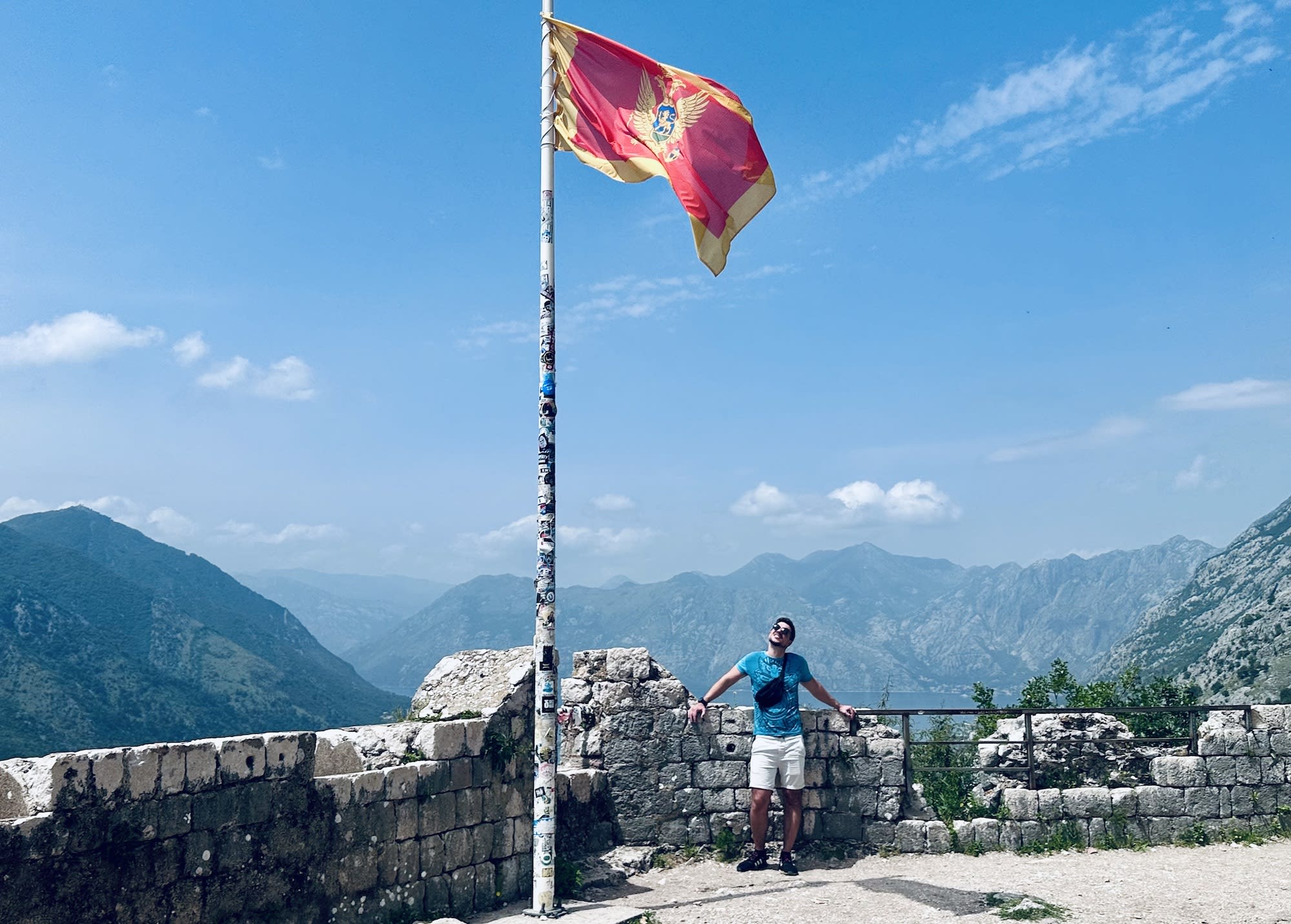 A person stands beside the Montenegro flag at the top of Kotor Fortress, with breathtaking mountain views in the background. Climbing to the top is one of the adventurous things to do in Kotor.