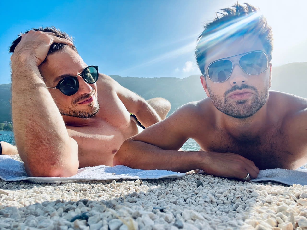 Two men relaxing on a pebble beach in Kotor under the bright sun, with mountains in the background. Enjoying the beach is one of the leisurely things to do in Kotor.