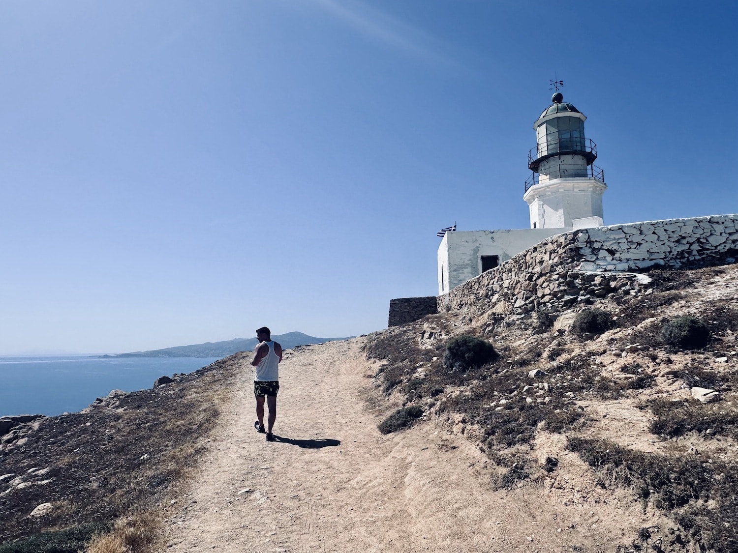 A person walks toward a scenic lighthouse on a rocky hill with the sea in the background, a perfect example of free and picturesque activities in Mykonos on a budget.