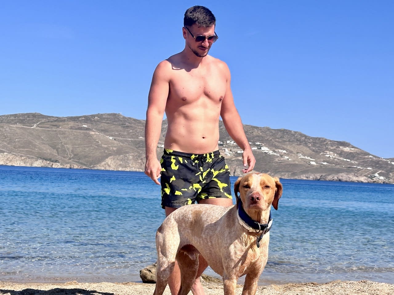 A man in black and yellow shorts walks his dog along a beautiful beach with clear blue waters and rugged hills in the background, highlighting an affordable and enjoyable activity in Mykonos on a budget.