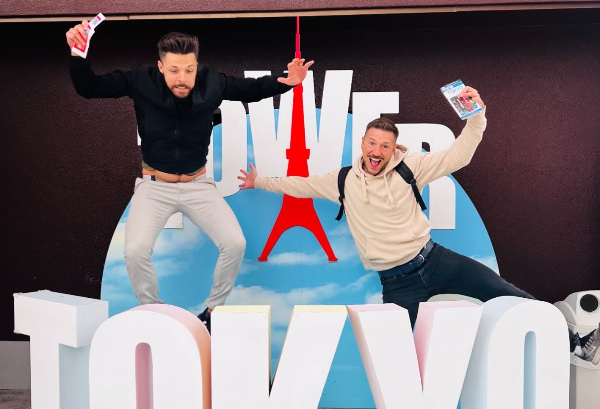 Two men are joyfully jumping in front of a sign that reads 'TOKYO,' with the iconic Tokyo Tower illustrated behind them, symbolizing the vibrant 2025 gay travel trends. The man on the left holds tickets, eagerly embracing the new wave of LGBTQ+ tourism anticipated in the coming years. Meanwhile, the man on the right extends his arms with a small item, perhaps a memento or guidebook, embodying the essence of adventure and cultural exploration that awaits queer travelers in Japan. Their smiles capture not only their personal excitement but also a broader invitation to all LGBTQ+ individuals seeking welcoming destinations like Tokyo for unforgettable travel experiences.