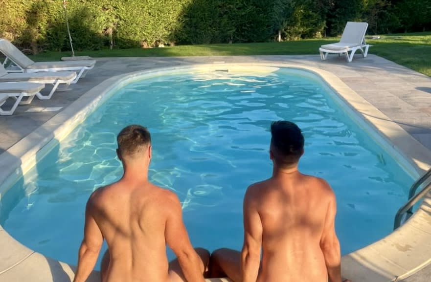 Two men sitting at the edge of a pool, facing away from the camera, in a private garden setting. They are attending a gay yoga retreat