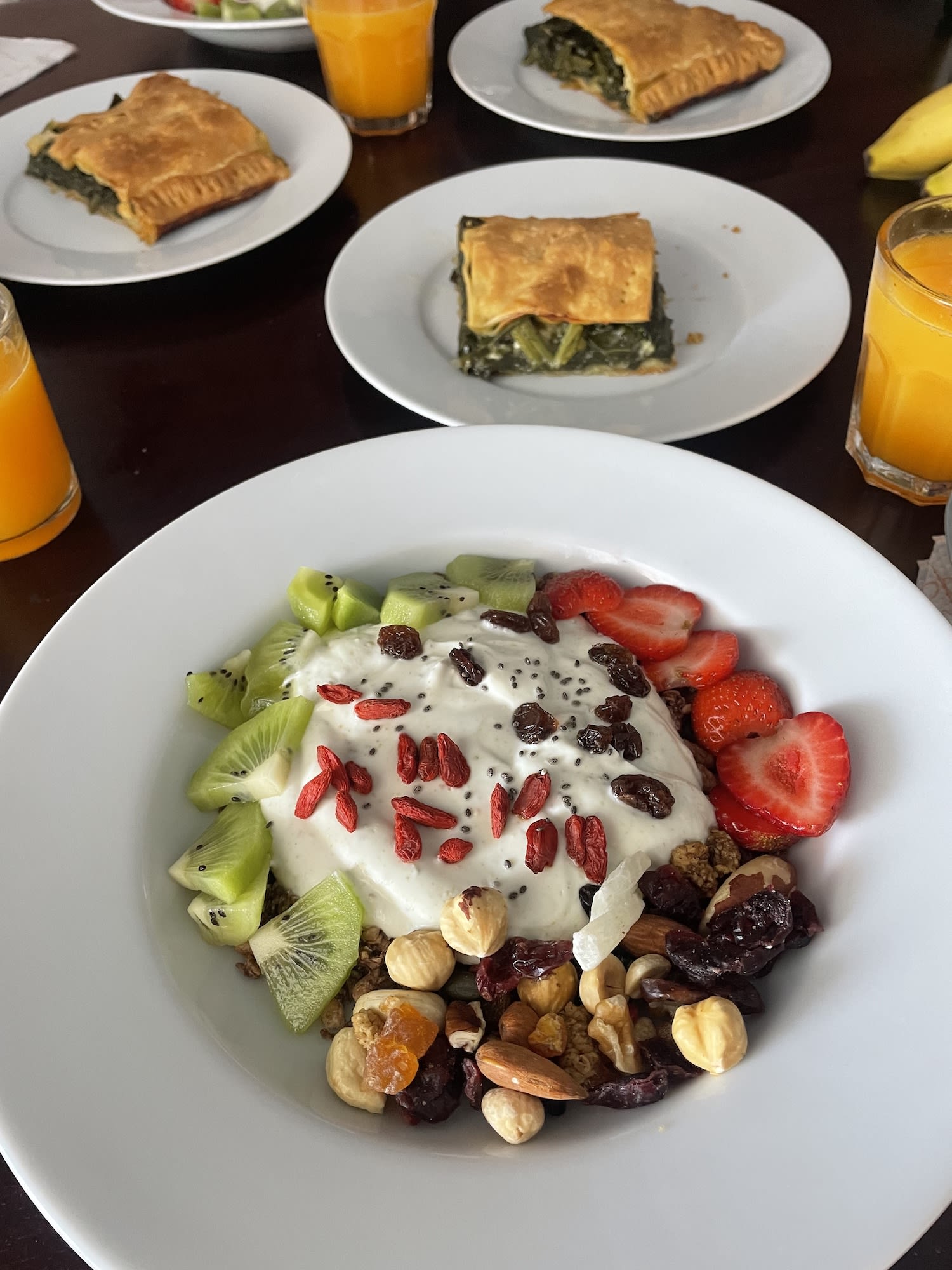 A bowl of yogurt topped with fruits, nuts, and seeds sits invitingly on the table. In the background, plates with spinach pastry and glasses of orange juice complement this delightful wellness-inspired breakfast scene.