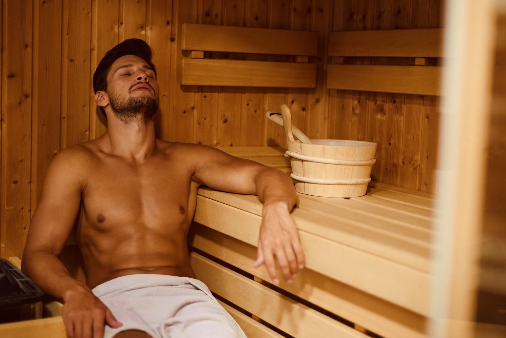 A relaxed man lounging in a wooden sauna, eyes closed, soaking in the heat, with a bucket and ladle beside him.