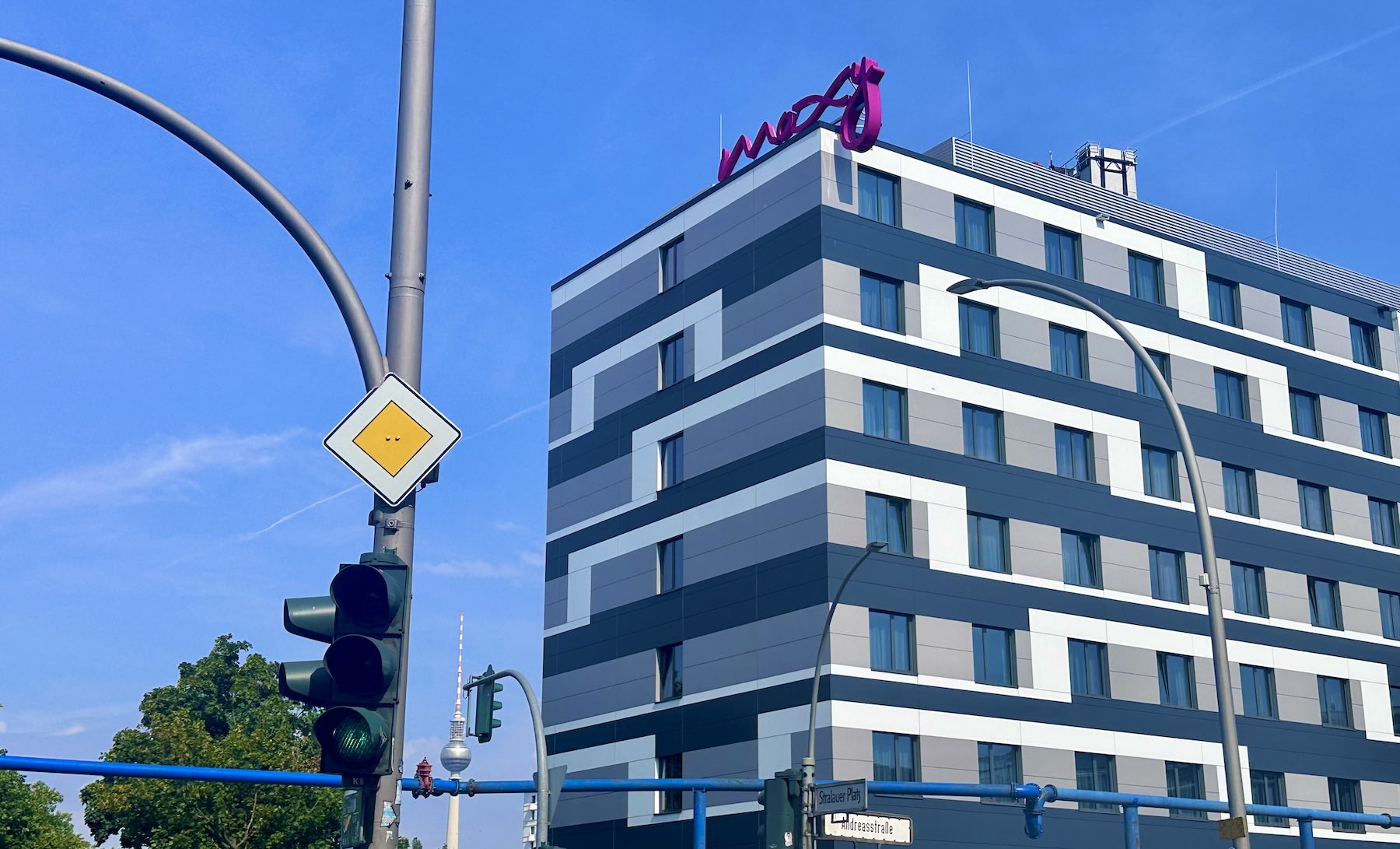 A modern multi-story hotel building with geometric patterns showcases its brand name in red on the roof, situated near traffic lights and street signs under a clear blue sky. Known for embracing diversity, this Berlin gem offers an inclusive stay and celebrates its vibrant community spirit, making it a standout choice among gay hotels in Berlin.
