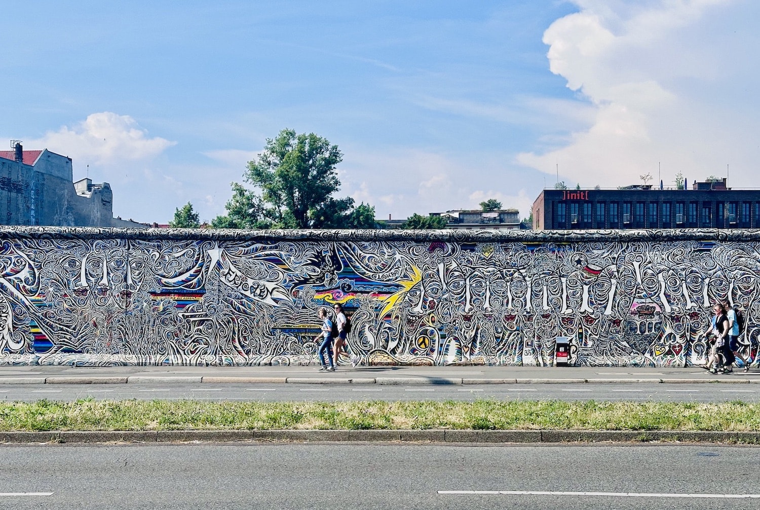 A colorful mural on a long wall in Berlin showcases abstract designs and intricate patterns celebrating diversity, echoing the inclusive spirit of gay hotels in Berlin. On this sunny day, passersby admire the vibrant artistry that enlivens the cityscape, reflecting Berlin's cultural richness and welcoming atmosphere for all travelers.