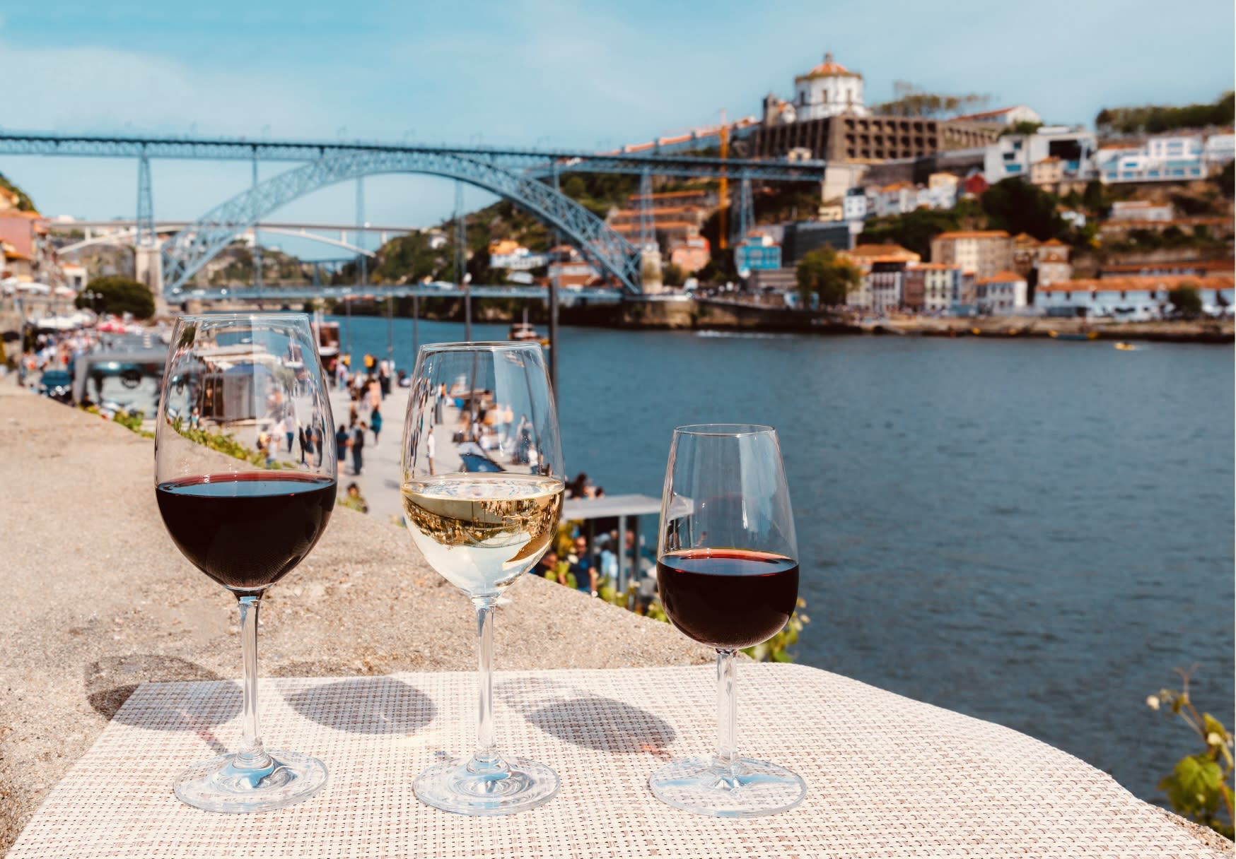 Three glasses of wine (red, white, and red) set on a table with a stunning view of the Ponte Dom Luís I bridge and the Douro River, showcasing the vibrant wine culture in gay Porto.