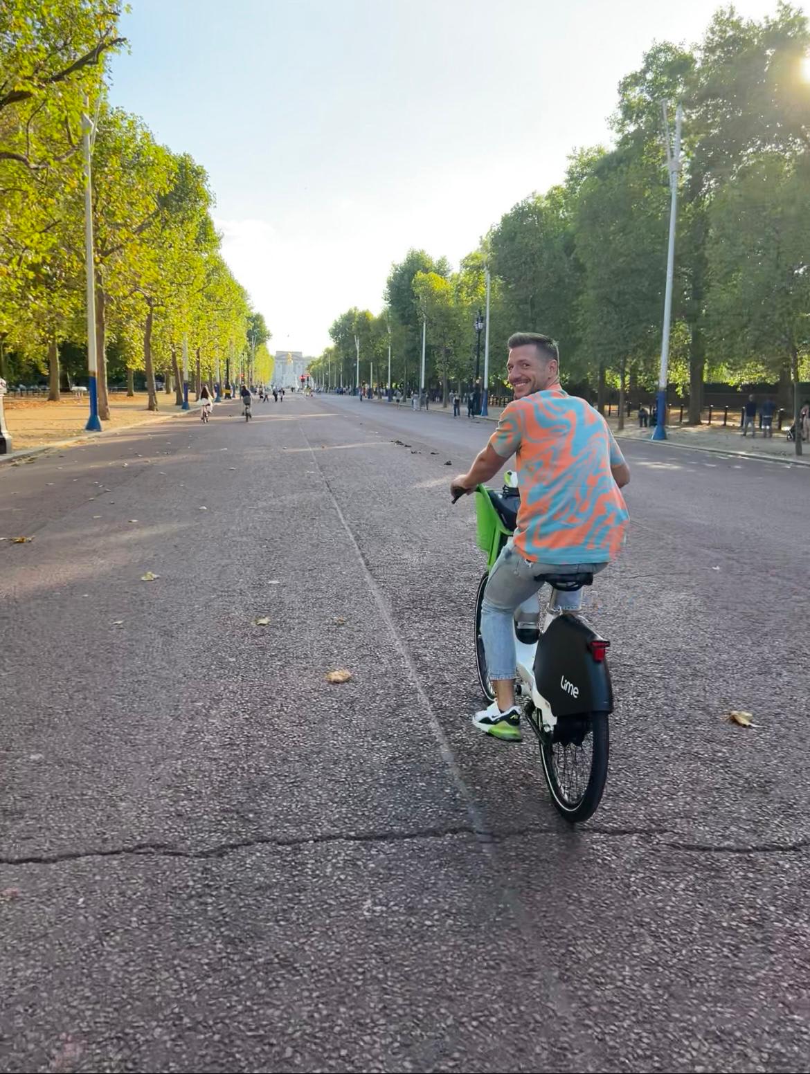 A man in a colorful tank top rides a Lime e-bike on an empty street lined with trees, smiling back at the camera. The bright and open setting, along with his cheerful expression, can brighten your day.