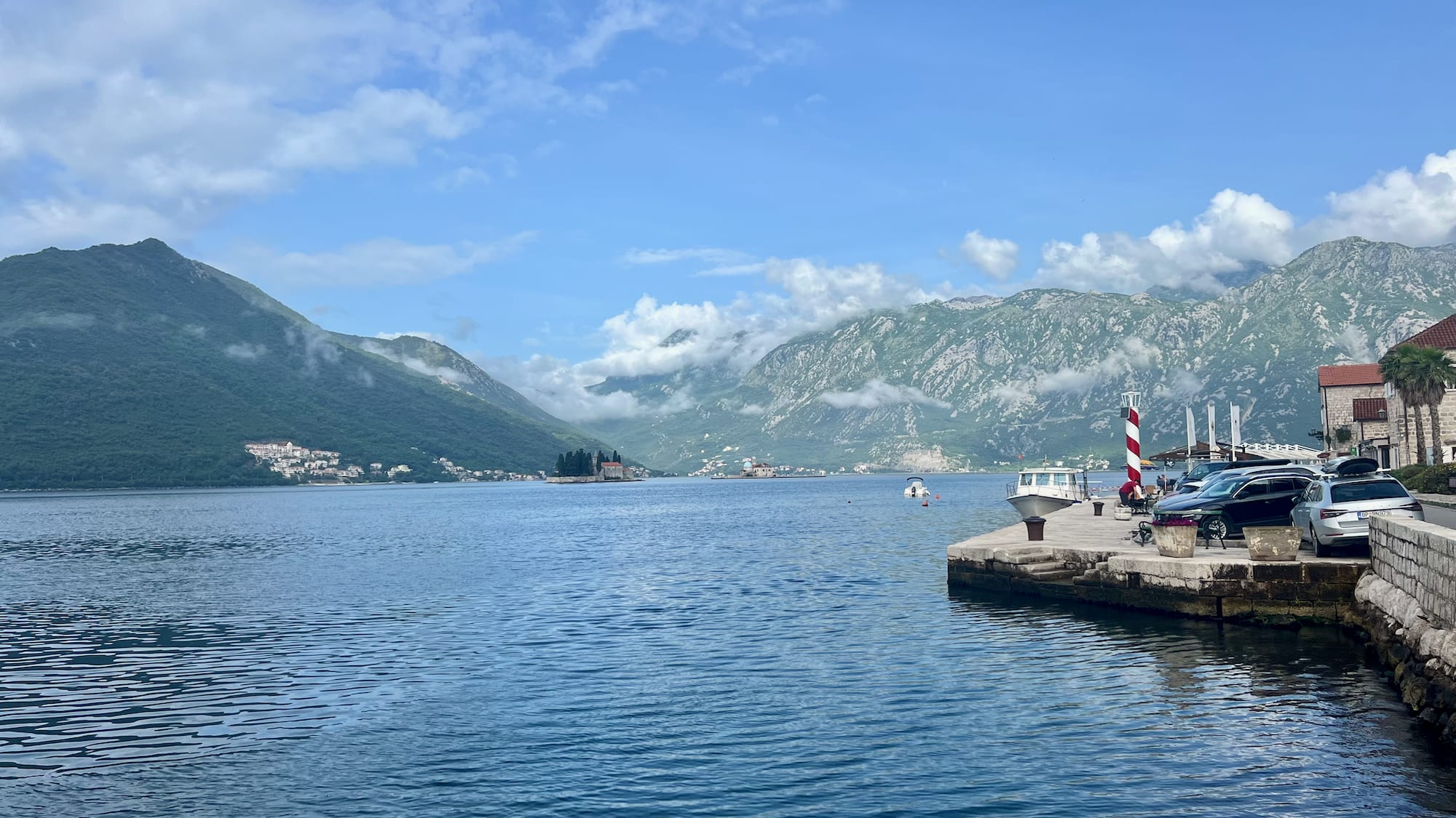 A scenic view of a calm bay with mountains captures the essence of a perfect Montenegro itinerary. On the right, a small dock features a red and white lighthouse, bustling with people and boats. This scene invites you to explore charming coastal towns nearby, making it an ideal stop on your Montenegro itinerary for those seeking natural beauty and local culture.