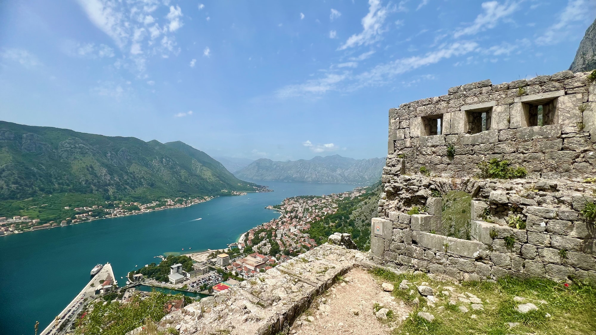 Explore the scenic view of a picturesque Balkan adventure with this Montenegro itinerary. A coastal town by the bay is highlighted, featuring a stone fortress standing proudly in the foreground. Majestic mountains serve as the perfect backdrop, creating an unforgettable scene for your travel journey.