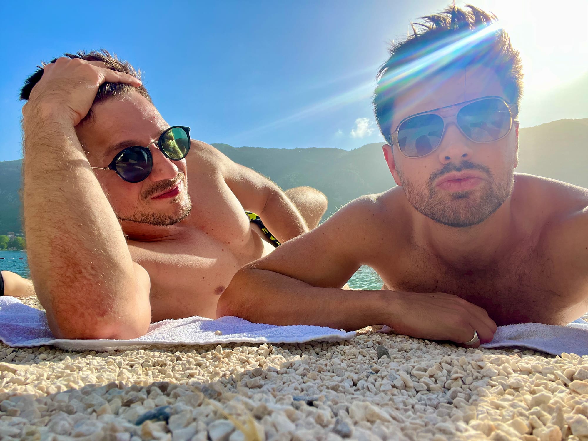 Two people in sunglasses relax on a rocky shoreline, soaking up the sun during their stunning Montenegro itinerary. The scene highlights mountains and sparkling water as a breathtaking backdrop to this Balkan adventure, capturing the essence of a perfect getaway in nature's embrace.