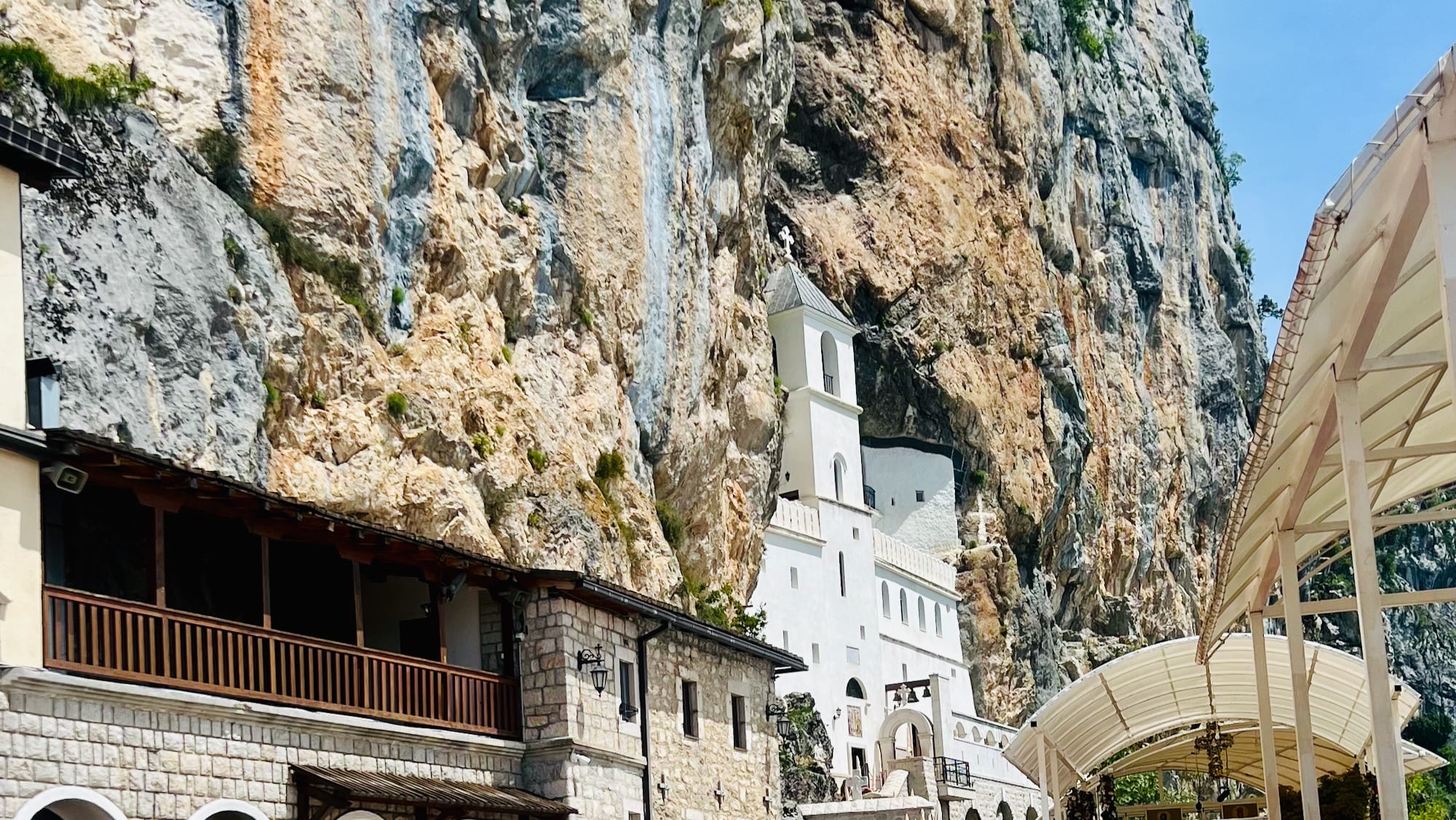Nestled into a rocky cliff, this monastery features white structures and arched stone buildings. A covered walkway can be seen in the foreground, offering a unique vantage point of this spiritual site—a must-see on any Montenegro itinerary or Balkan adventure.
