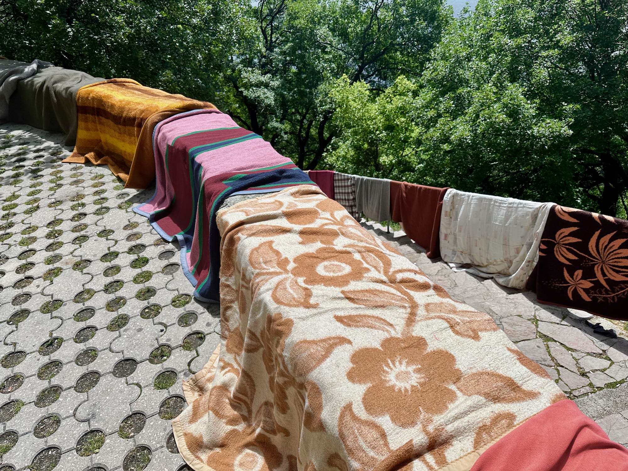 Colorful blankets and towels hang on a sunlit balcony railing, with lush green trees in the background. This serene scene captures a relaxing moment amidst your Montenegro itinerary, offering a glimpse into local life and nature's beauty.