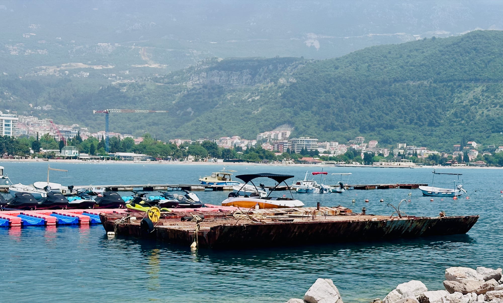 A serene Montenegro itinerary scene captures boats and a floating platform on tranquil waters, with majestic mountains rising behind charming coastal towns.