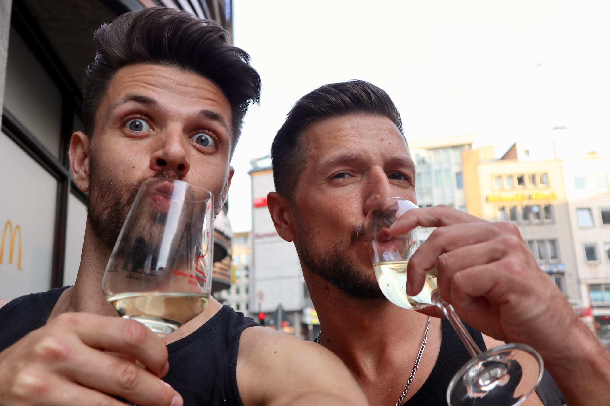 Two men, smiling broadly, take a selfie as they clink glasses of white wine, embodying the lively spirit of LGBTQ+ nightlife. The scent of gay cologne subtly lingers in the vibrant urban air, adding an aromatic layer to their joyful celebration. Bright city lights twinkle around them, enhancing the scene's dynamic energy and sense of community pride.