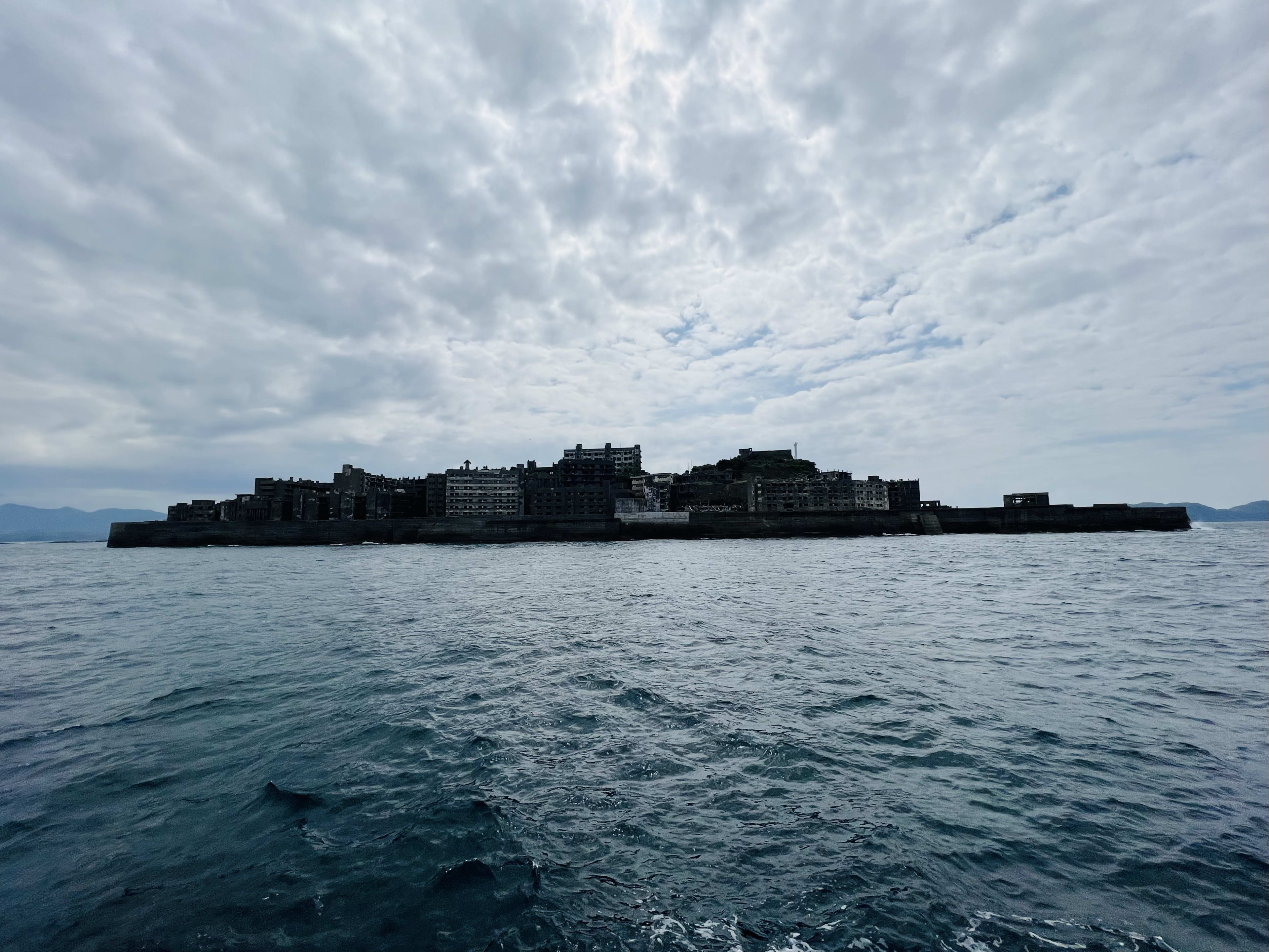 A distant view of an abandoned island, its cluster of dilapidated buildings standing stark against the sea under a cloudy sky—Hashima Island (Gunkanjima), a fascinating destination to include on your japan itinerary, especially while exploring Kyushu before heading north.