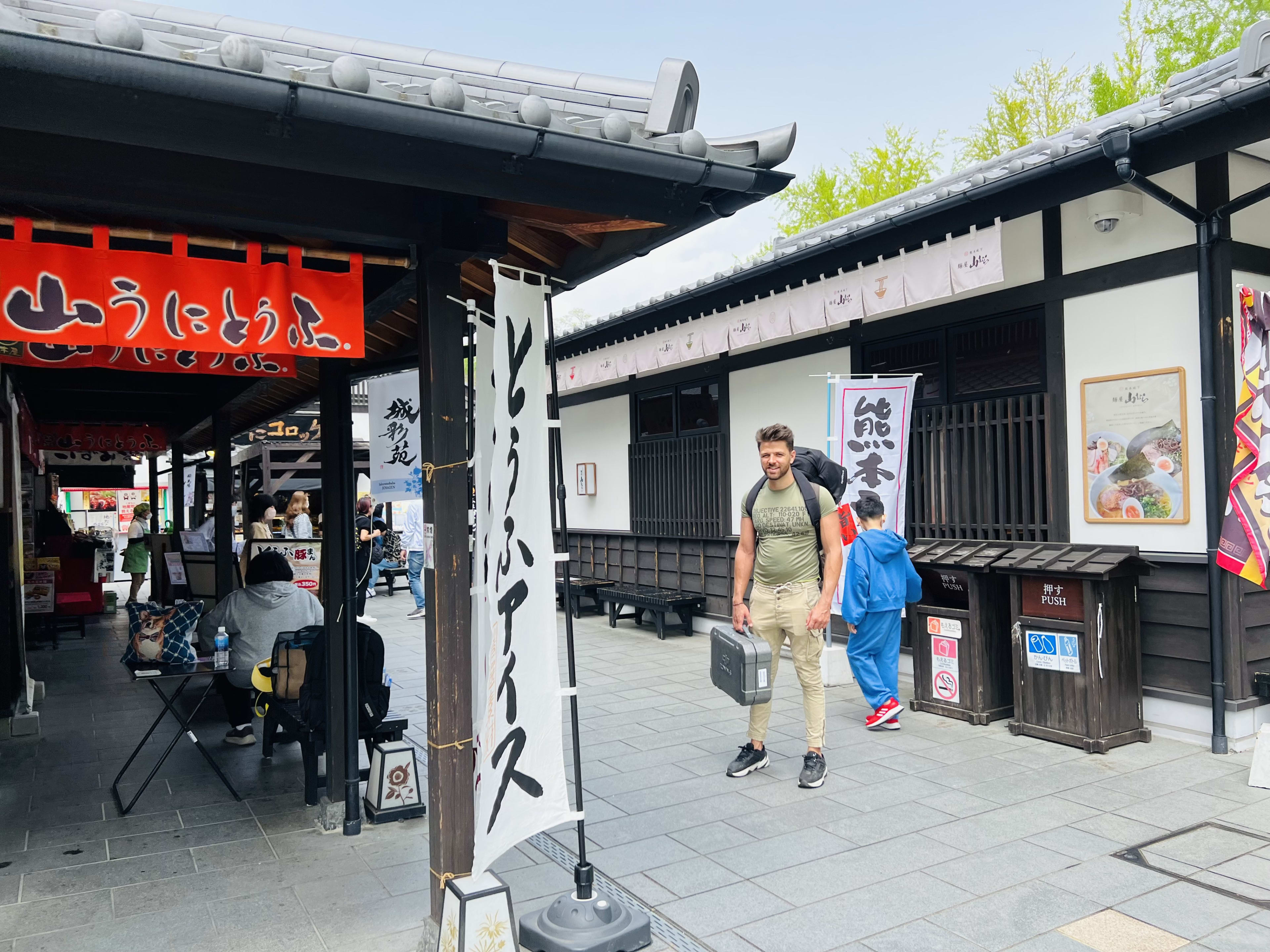 A man carrying a suitcase leisurely explores Kyushu, strolling through a traditional Japanese shopping street lined with wooden buildings. People sit at a cafe on the left, and colorful banners hang from the eaves under a clear sky.