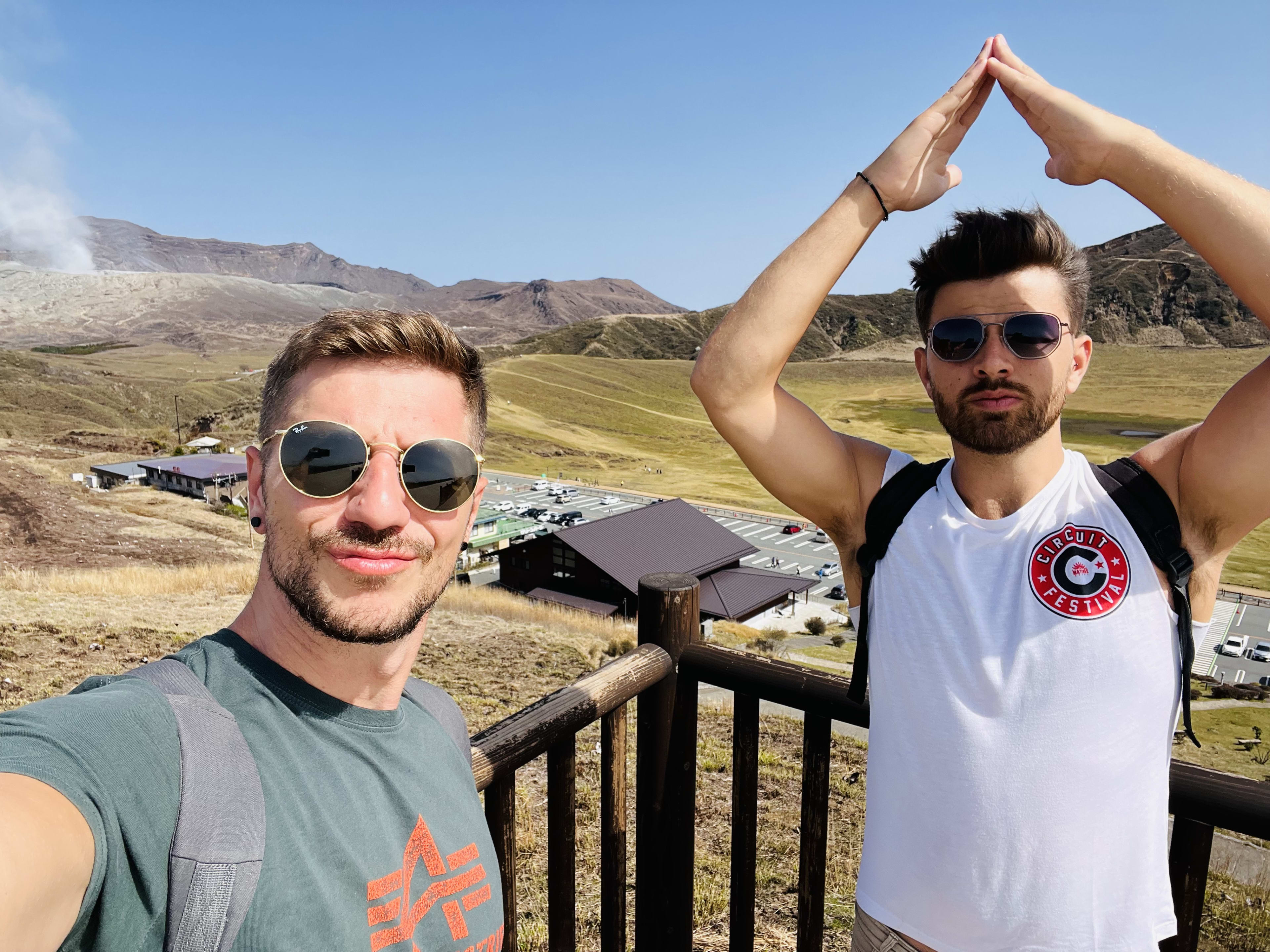 Two men wearing sunglasses take a selfie at a scenic overlook, with green mountains and a traditional building in the background. One man raises his arms above his head, celebrating their travel moment. The photo captures the joy and highlights a stop on their japan itinerary during their adventure.