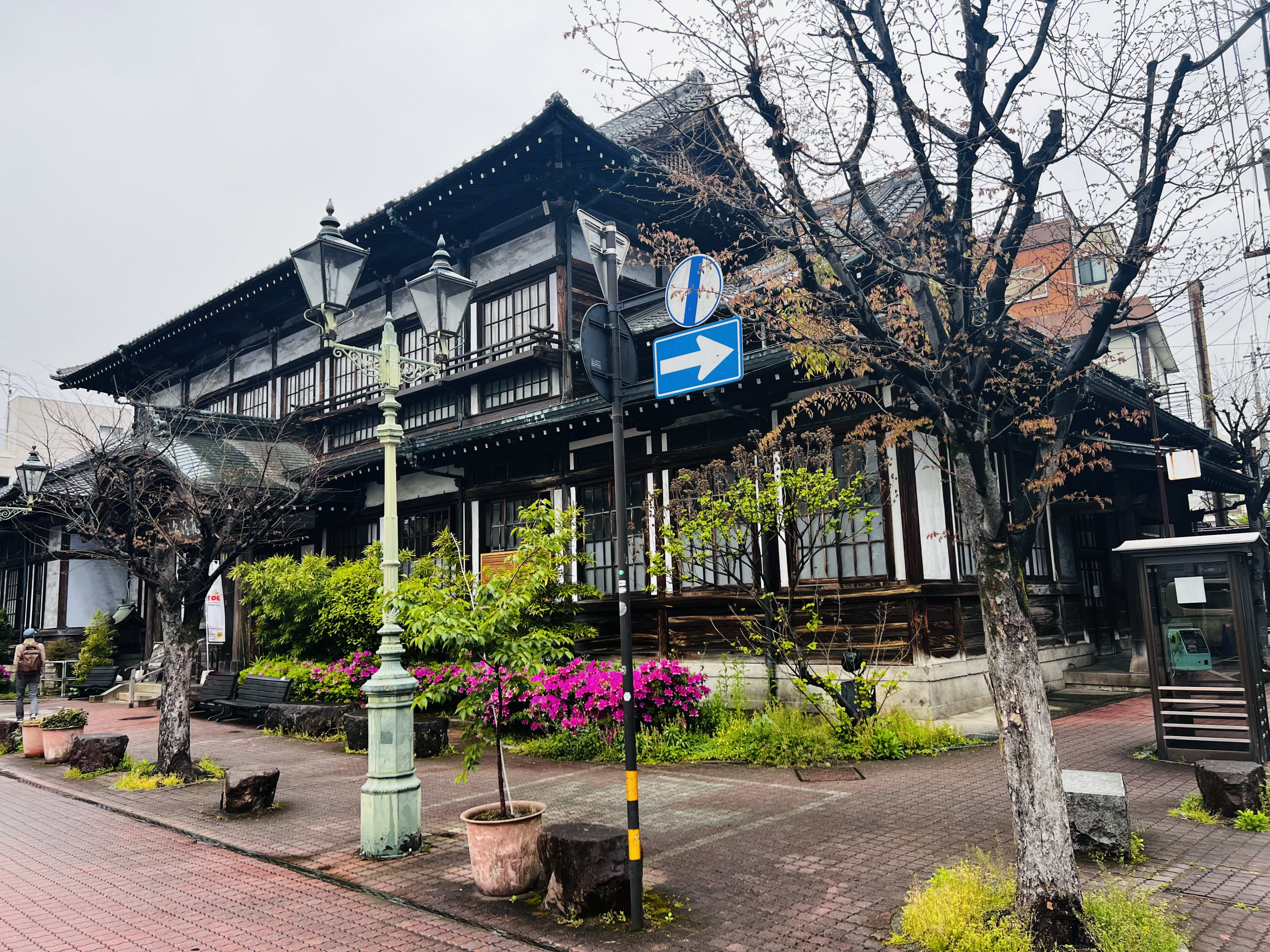 A traditional Japanese-style building with dark wood accents and white walls graces the street, inviting you to explore Kyushu. The scene is adorned with bare trees, vibrant pink flowers, and a vintage lamp post. A blue traffic sign points right, guiding travelers through the wonders of Kyushu travel.