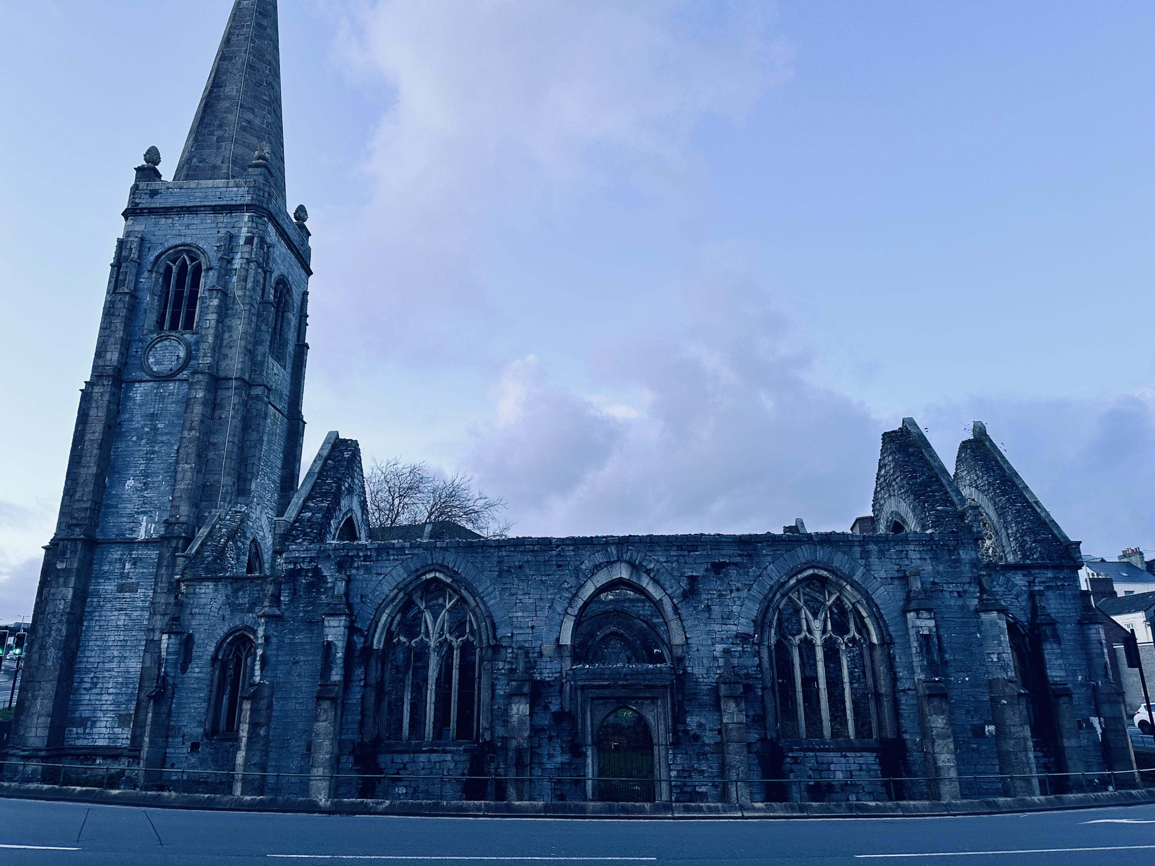 Twilight view of the Charles Church, a historical landmark in Plymouth, with its stone gothic ruins and spire standing against a dusky blue sky.