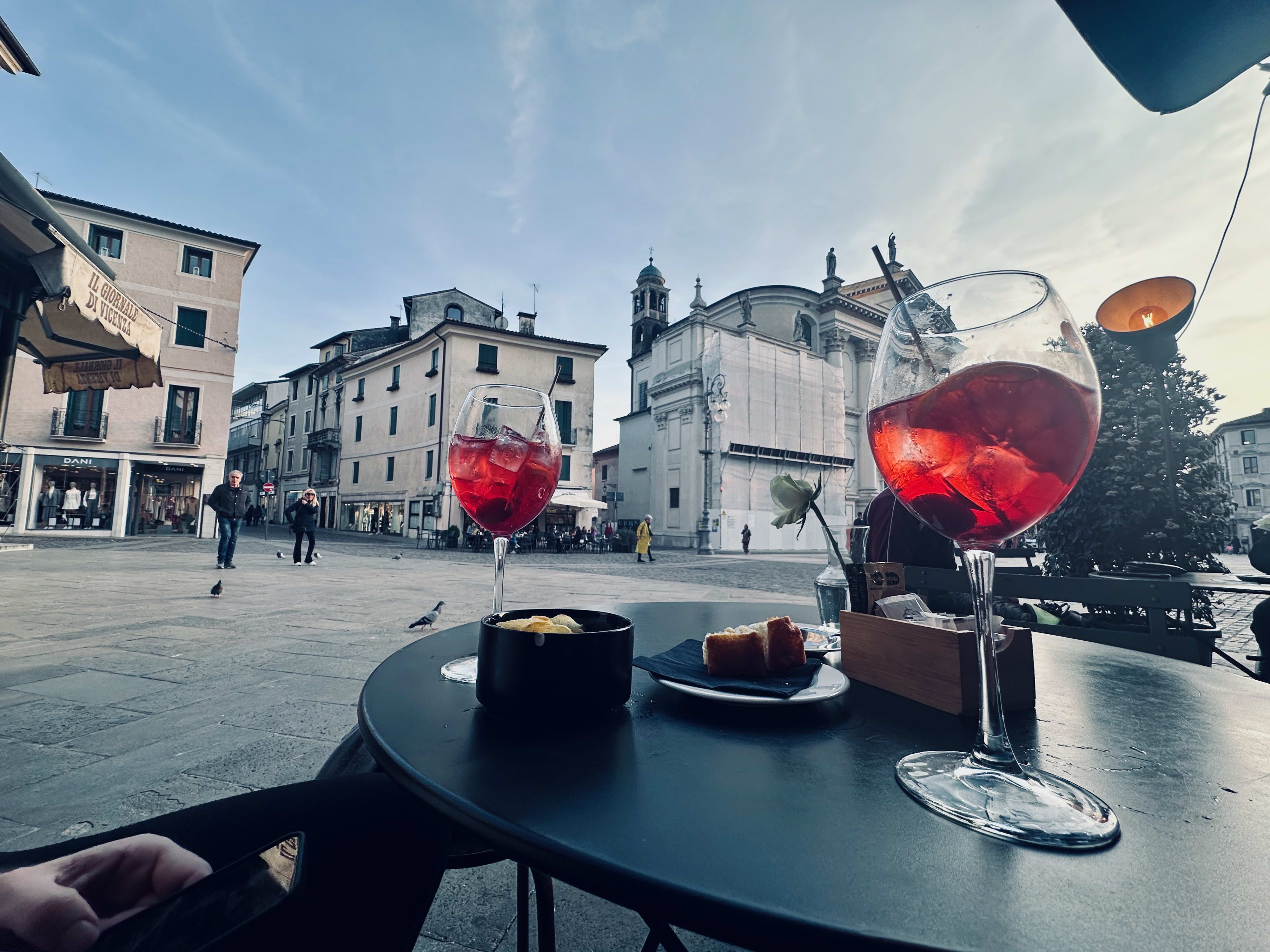 Two glasses of Campari Spritz adorn a cafe table, perfectly paired with traditional snacks in the heart of a European plaza. Historic buildings frame the scene while locals and tourists alike soak in the atmosphere. This moment captures the essence of relaxation during your 2 day Venice itinerary, offering a taste of local culture amidst stunning architecture and vibrant street life.
