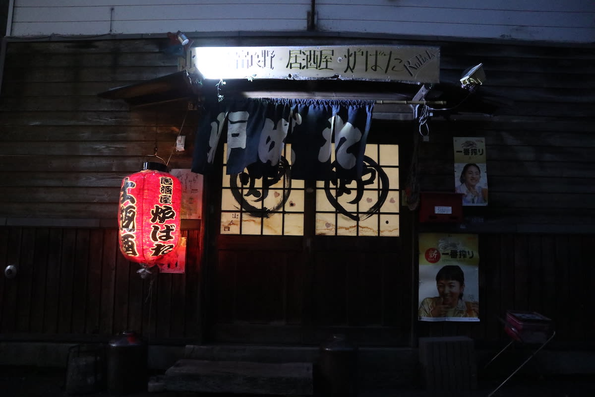 A cozy, dimly lit wooden storefront features a glowing red paper lantern and traditional blue noren curtains, with colorful posters beside the entrance—an inviting scene you could encounter along your 3 week Japan itinerary.