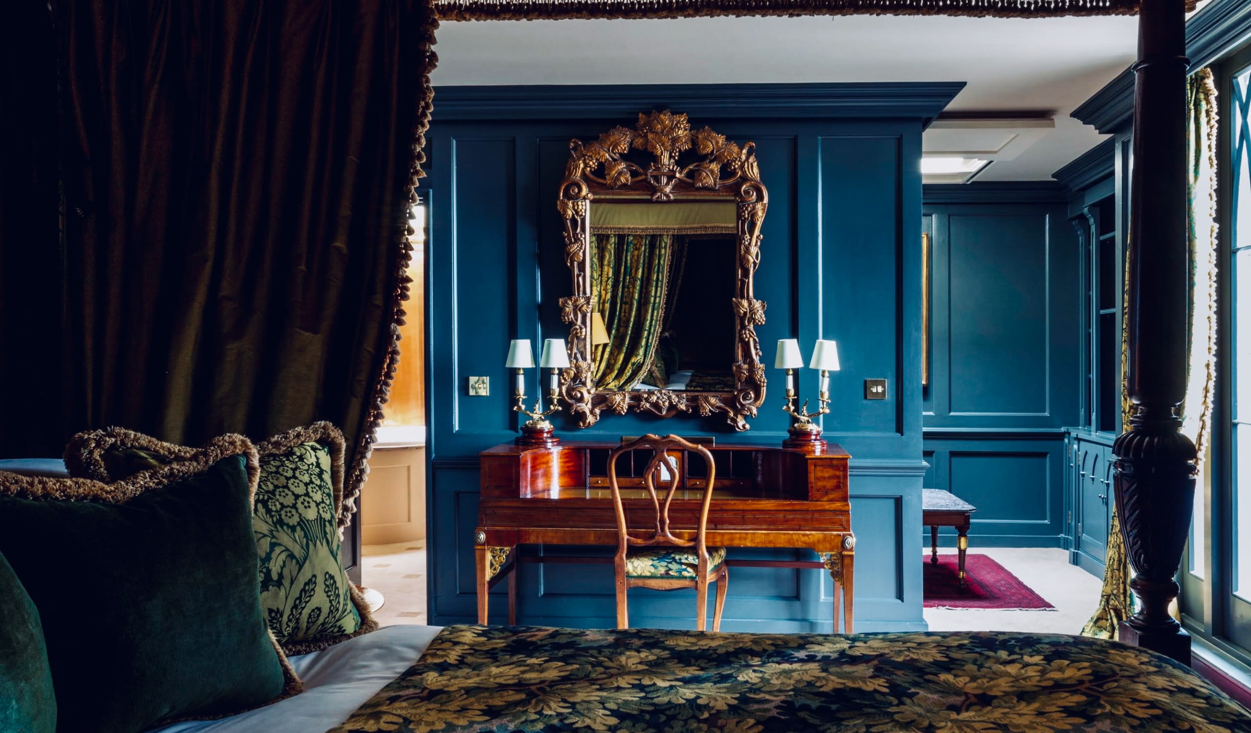 Luxurious bedroom in one of the finest gay London hotels, showcasing ornate decor with a spacious mirror, elegant wooden desk, and comfortable upholstered chair. The bed is beautifully adorned with patterned pillows. The inviting ambiance is enhanced by dark blue walls and intricate molding, embodying the sophisticated charm of an exclusive retreat for travelers in London.