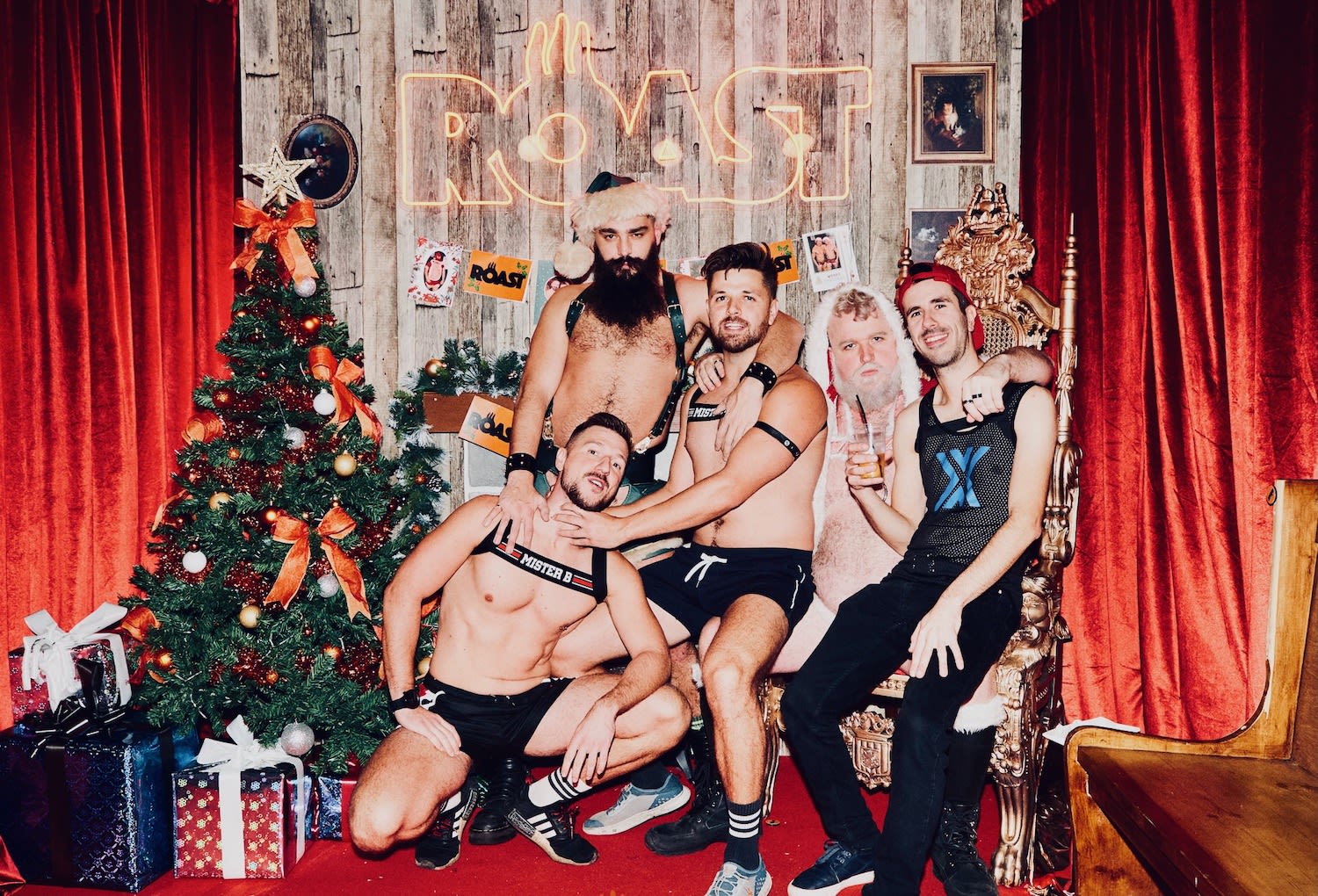 Four men in festive attire pose cheerfully by a Christmas tree and gifts, beneath a vibrant neon sign. They stand on a stage adorned with red curtains, embodying the joyful spirit found in gay-friendly holiday destinations, where inclusivity shines bright.