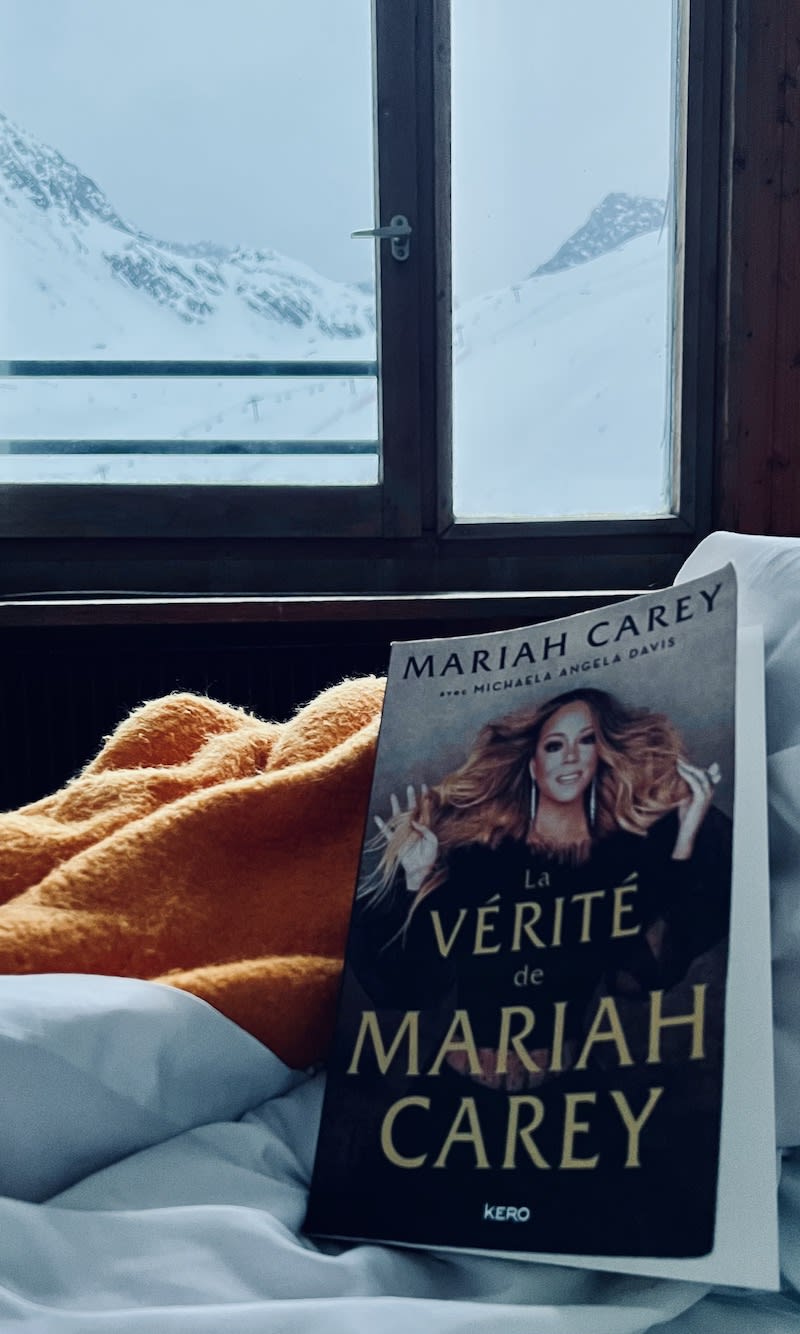 A cozy ski lodge room with a view of snow-covered mountains through a window, and a Mariah Carey autobiography on the bed, suggesting a relaxing read during Gay Ski Week.