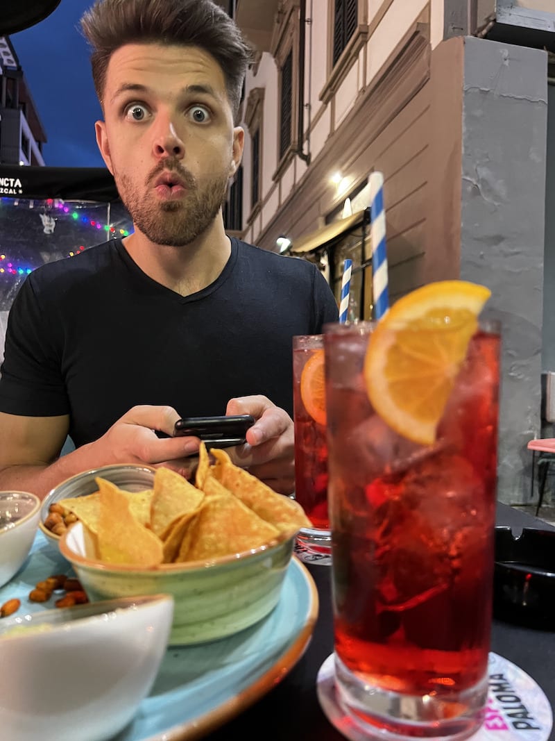 A surprised man with a whimsical expression sitting at an outdoor table, with a close-up of a red cocktail adorned with a lemon slice and a plate of chips and guacamole, capturing a lively aperitivo scene in Milan.