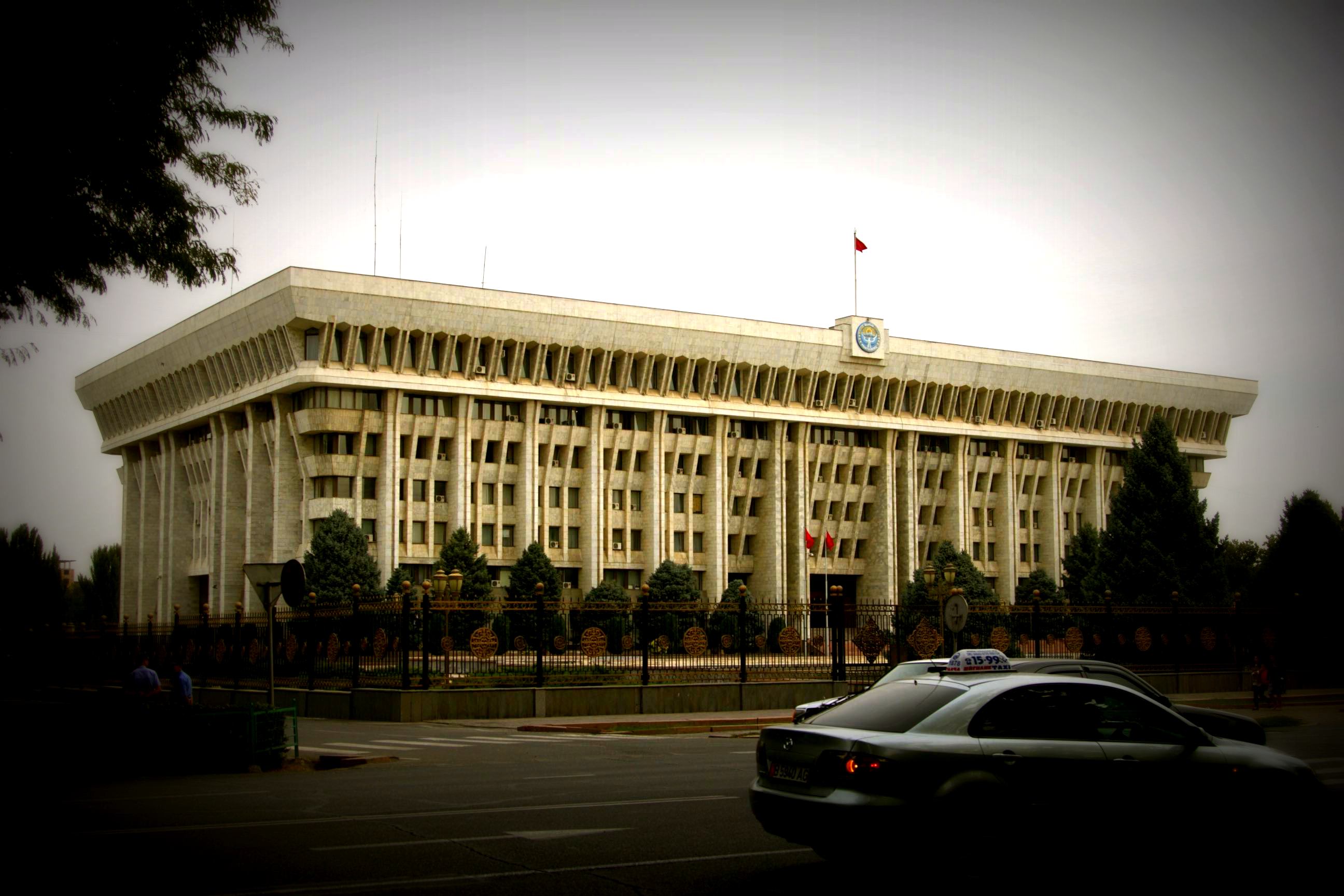 The imposing facade of the House of Government in Bishkek, Kyrgyzstan, framed by ornate fencing and dark skies, a significant landmark in the city.