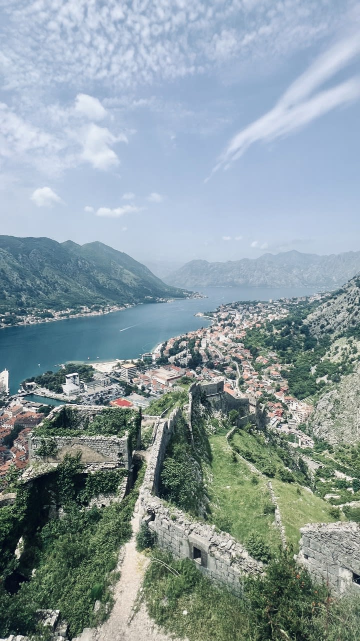 A panoramic view from the Kotor Fortress overlooking the Bay of Kotor, with ancient stone walls winding through lush greenery, and the historic town of Kotor spread along the shoreline. An essential activity when exploring things to do in Kotor.