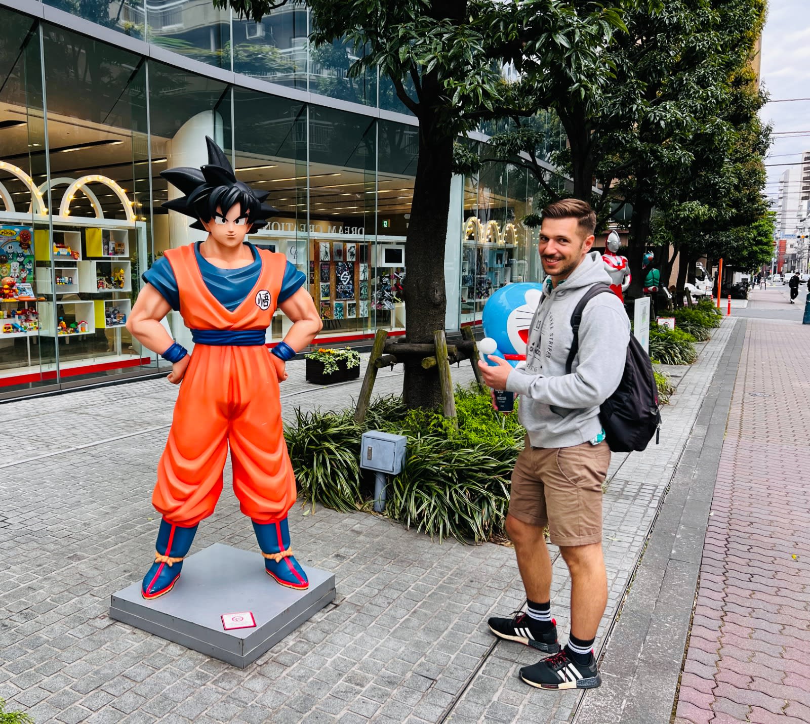 A man stands beside a large Goku statue on a bustling city sidewalk, holding a Doraemon figure with a colorful toy store in the background—an ideal stop for anime fans planning their japan itinerary. The scene captures the vibrant pop culture found throughout Japanese cities.