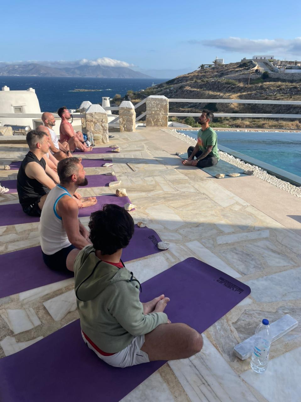 Group of men participating in a gay yoga retreat class on a terrace overlooking the sea, with mountains in the background.