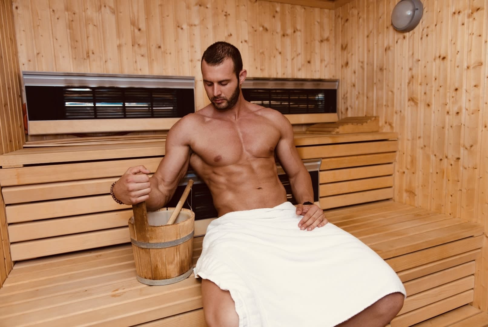 An attractive, muscular man sitting in a modern wooden sauna, holding a ladle over a bucket, wrapped in a towel, enjoying the warmth and ambiance.