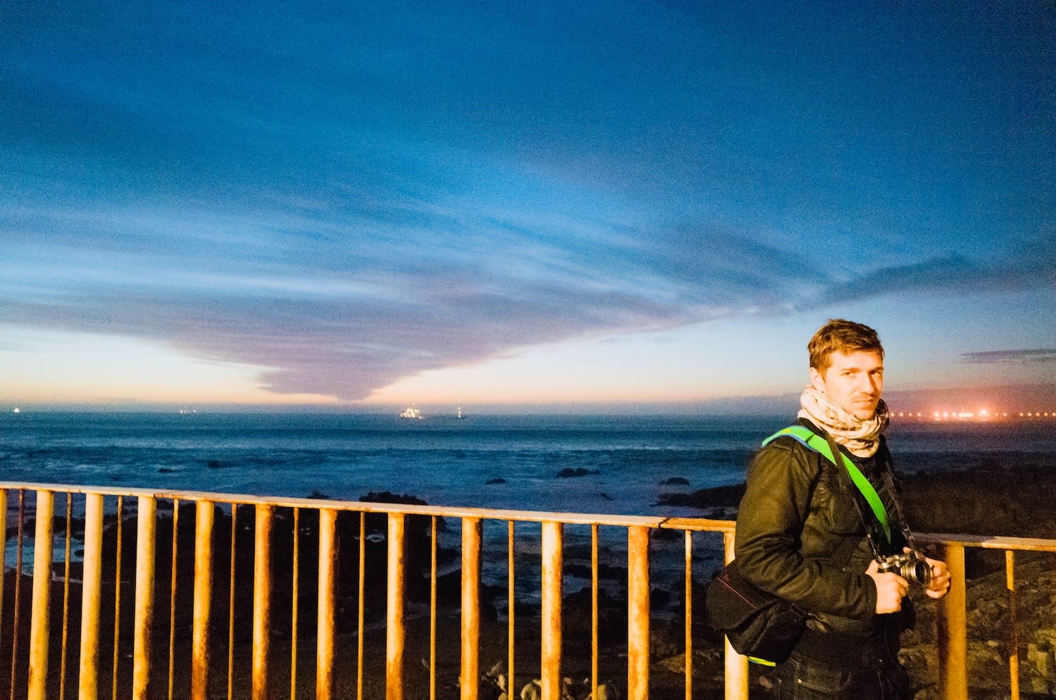 A man standing by a rusted metal railing overlooking the ocean at sunset, capturing the serene and picturesque coastal vibe of gay Porto.