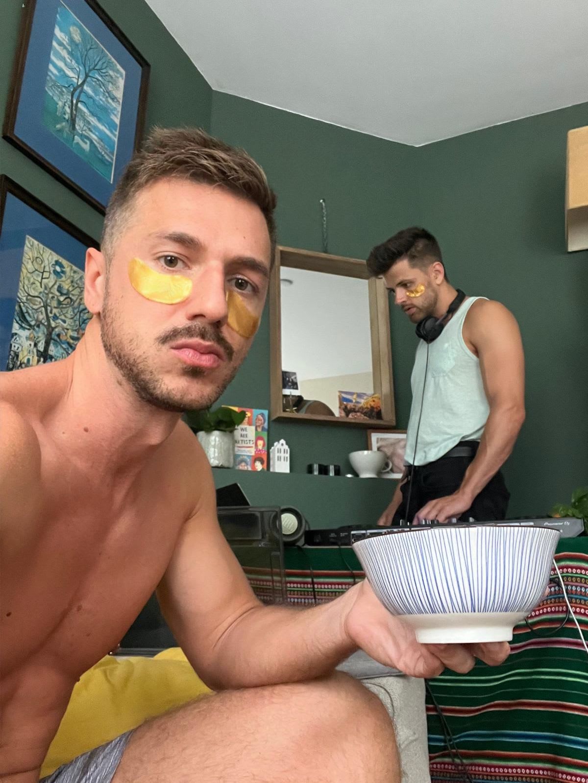 A man applies under-eye patches while holding a bowl, and another man in the background is DJing. This scene of self-care and hobbies at home can brighten your day with its relaxed vibe.