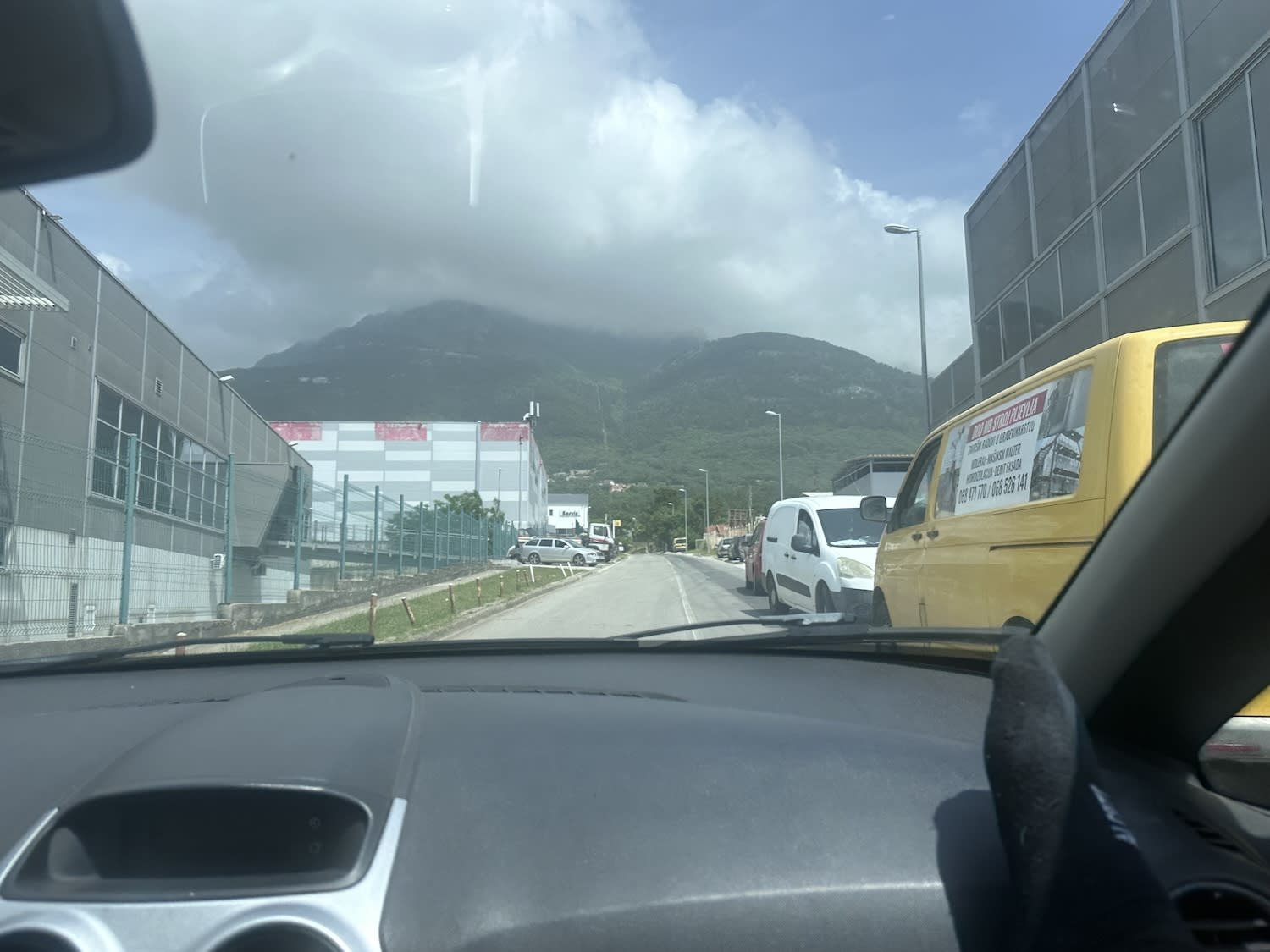 View from inside a car driving through an industrial area in Podgorica, Montenegro, with mountains in the background, highlighting a typical road scene for rental car users.
