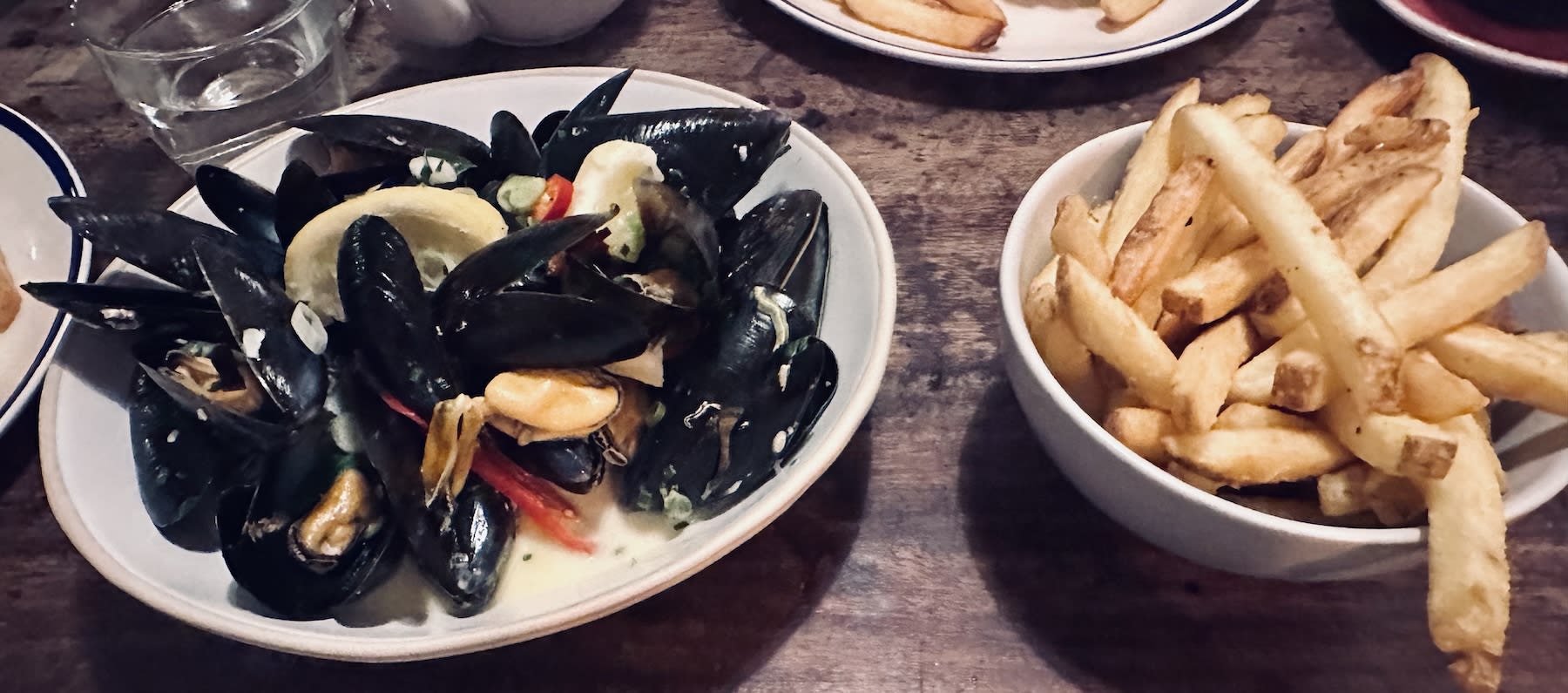 A plate of mussels in a creamy sauce with a lemon wedge, accompanied by a bowl of crispy fries, served at The Boot Factory in Bristol. A popular dining choice in the city.