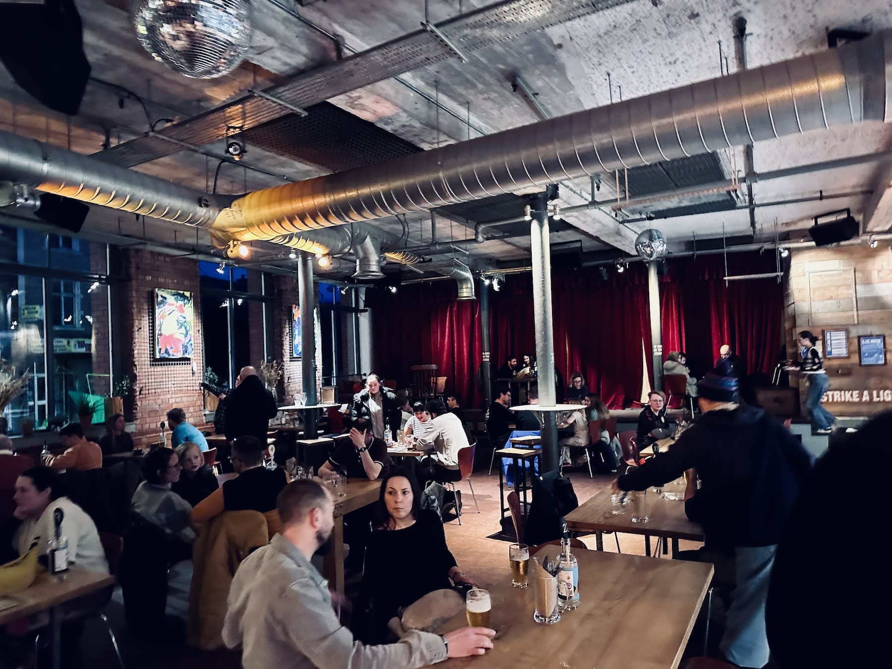 Inside the Tobacco Factory in Bristol, patrons are gathered in a spacious, industrial venue with exposed pipes and brick walls. The lively atmosphere is typical of the city’s social scene, often frequented by the LGBTQ+ community.