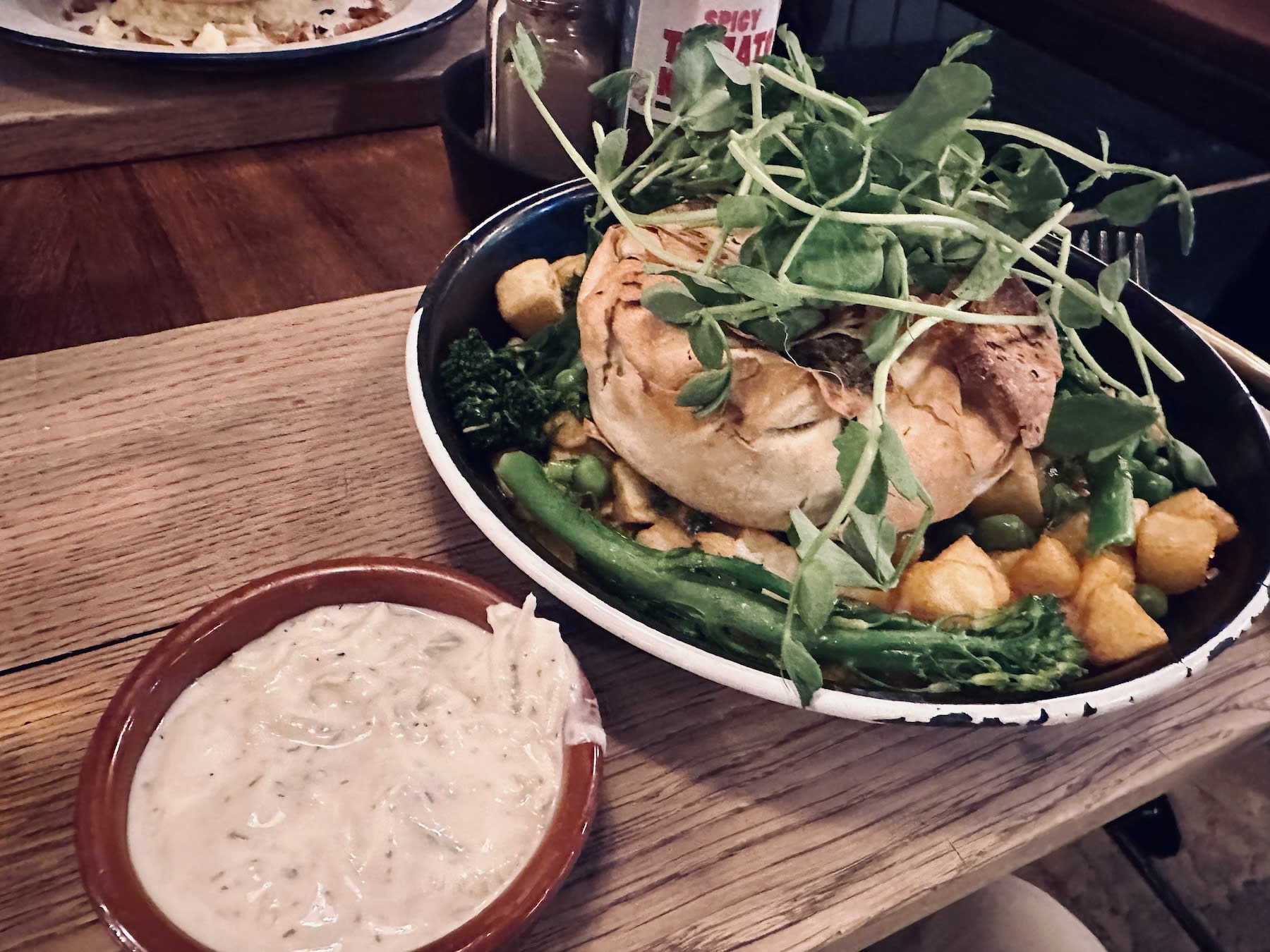 A savory pie topped with greens and served with roasted potatoes and tenderstem broccoli at Pieminister, a popular spot in Bristol. A small dish of creamy sauce is placed alongside, adding to the hearty British meal experience.