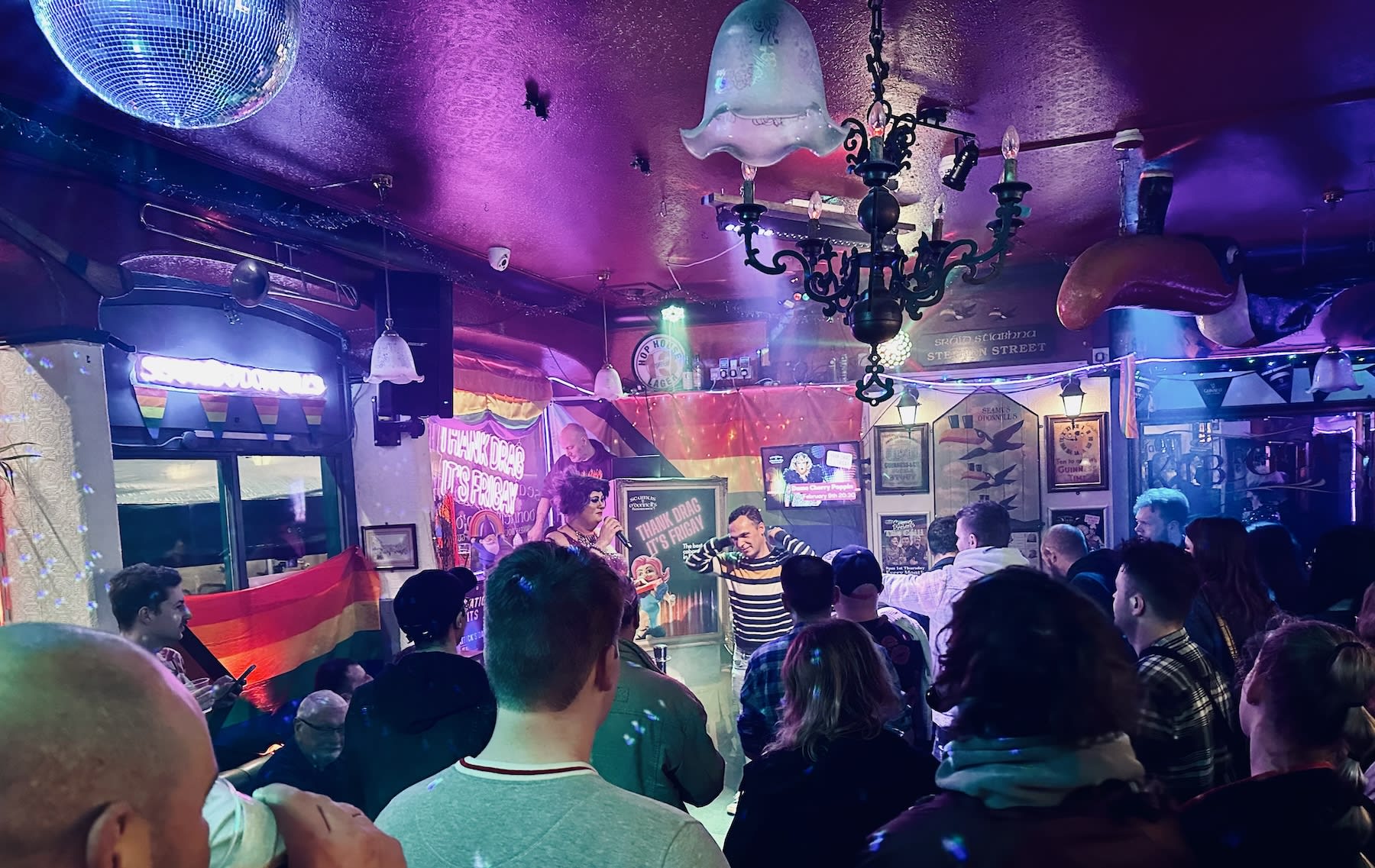 A vibrant drag show is underway at Seamus O’Donnell’s pub in Bristol, with a drag queen performing on stage under colorful lighting. The pub is filled with an enthusiastic crowd, reflecting the inclusive atmosphere.