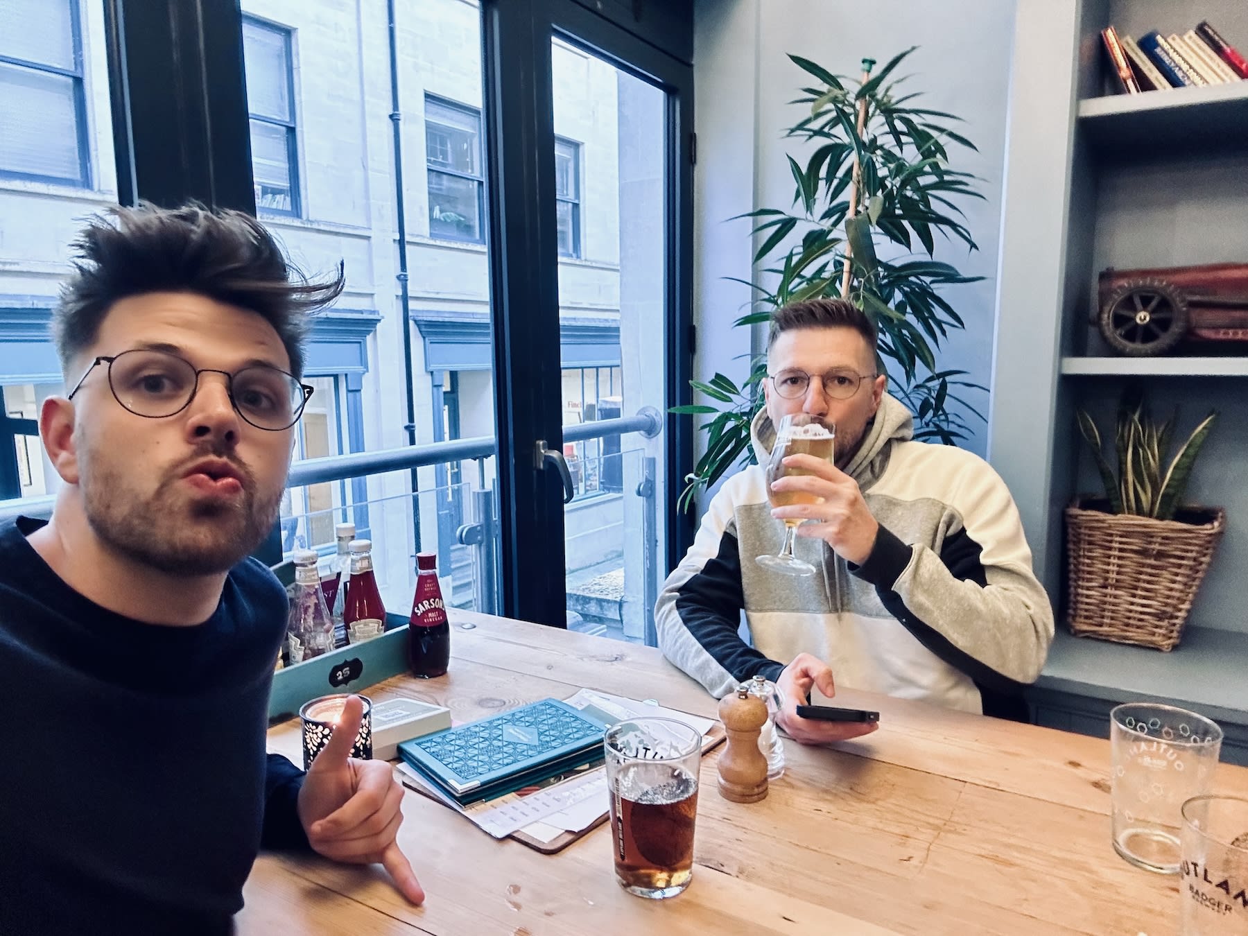 Two men enjoying drinks at a wooden table inside Hall & Woodhouse, a popular pub in Bath, United Kingdom. The cozy interior features large windows and modern decor, making it a favored spot for both locals and visitors, including those in the LGBTQ+ community.