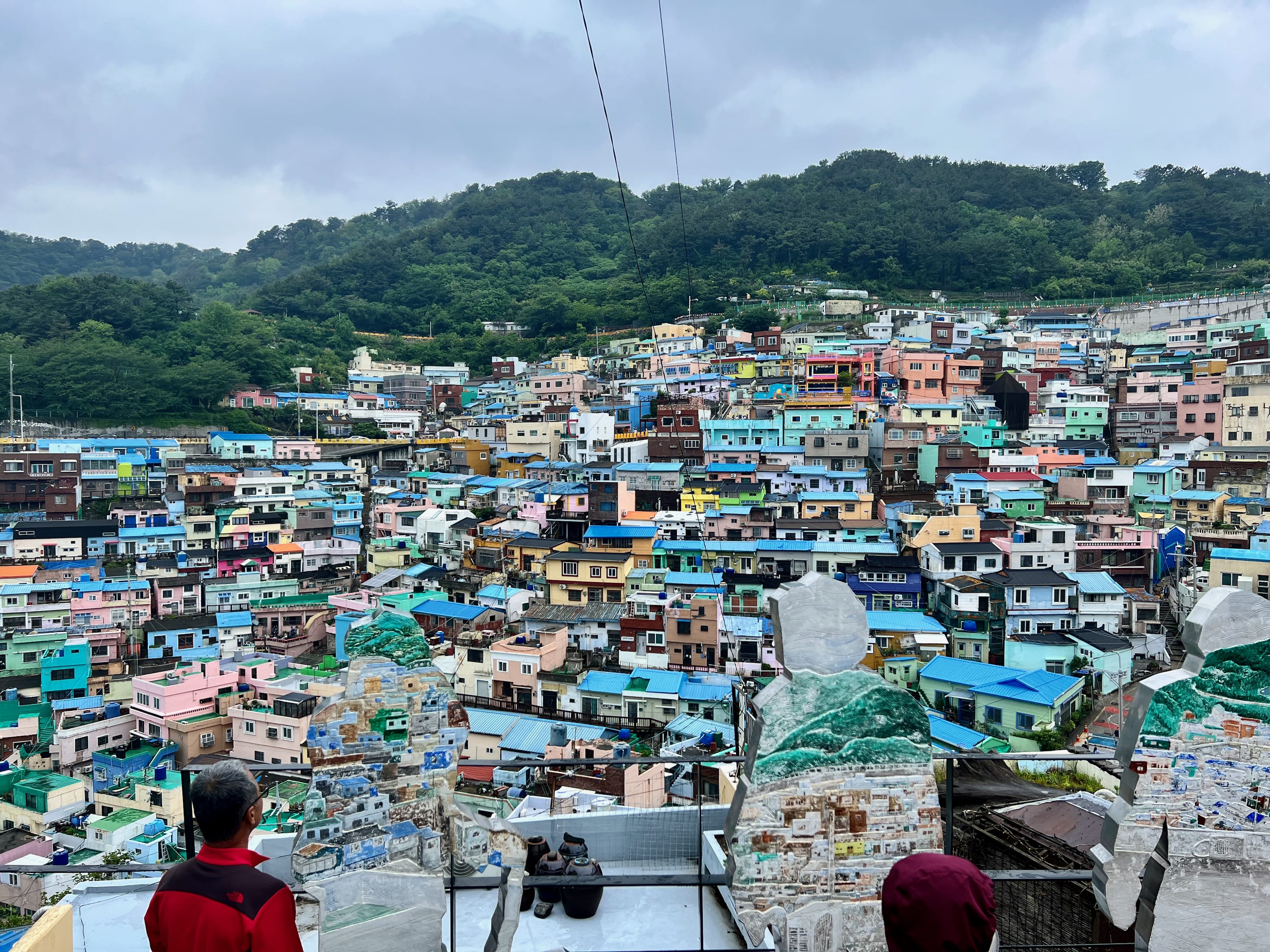 A vibrant hillside village in Busan features colorful, densely packed houses in shades of blue, teal, yellow, and pink. A lush, green forest and cloudy sky provide a backdrop. In the foreground, two people look over a stone wall at the scenic view of this enchanting coastal city.