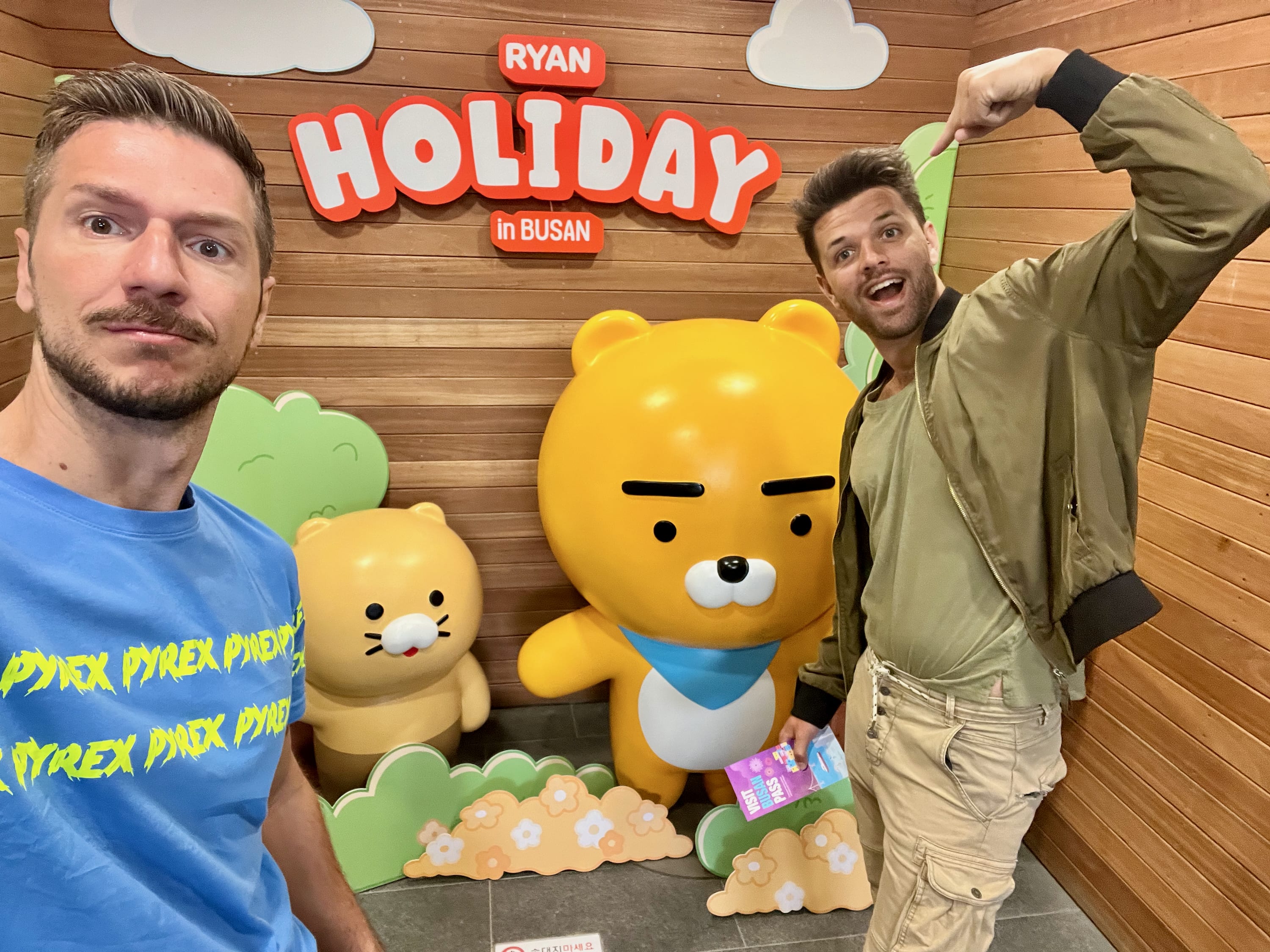 Two men pose energetically beside large cartoon bear statues, one smaller and one larger, wearing blue scarves. They stand in front of a wooden wall with 'RYAN HOLIDAY in BUSAN' boldly displayed, capturing the spirit of this vibrant coastal city. One wears a blue shirt with neon yellow text, the other a tan jacket.
