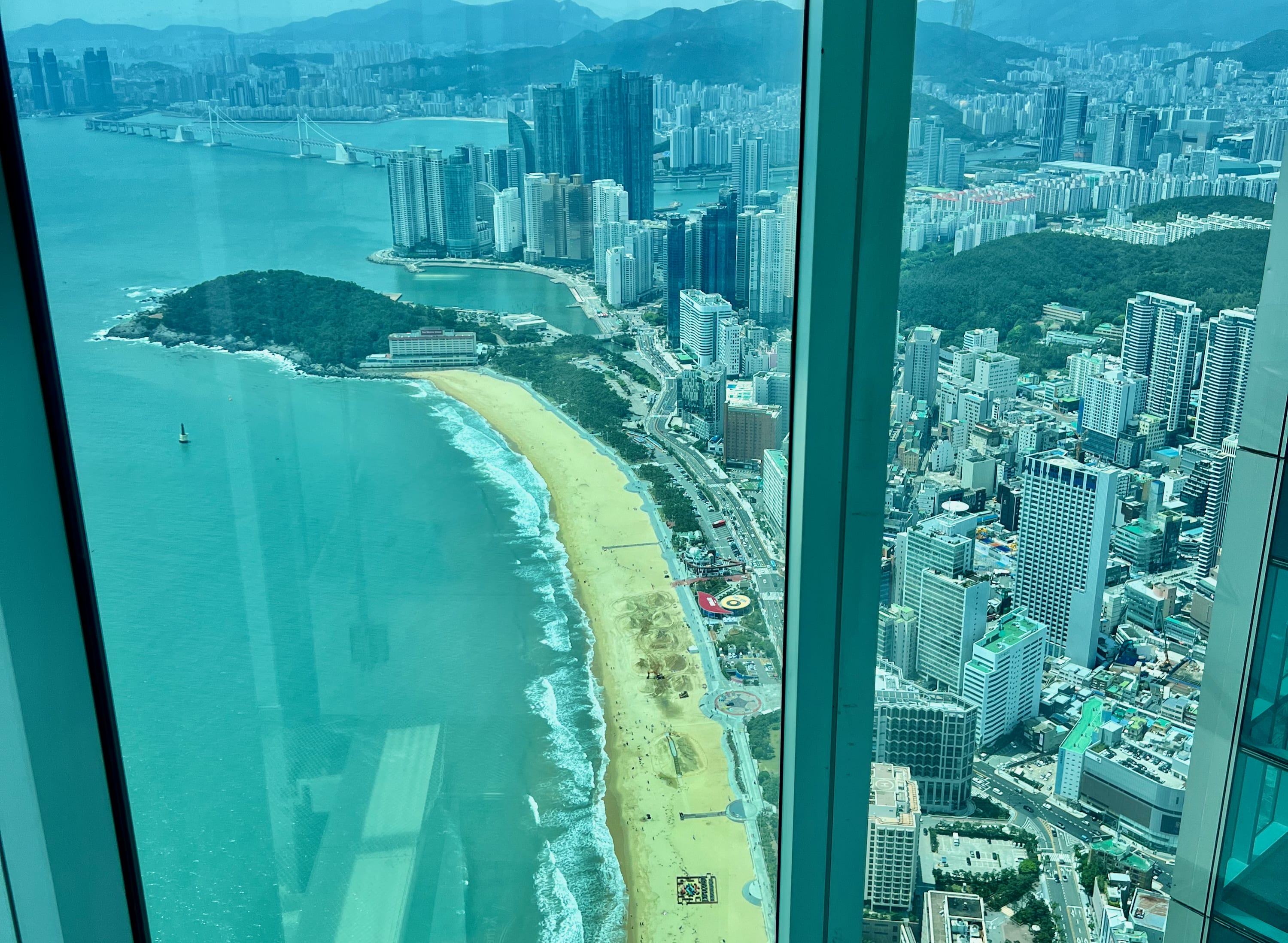 Aerial view through a window shows the sandy beaches of Busan, a vibrant coastal city by the sea. High-rise buildings and a curving shoreline are visible, with lush green hills in the background. A large bridge spans the water in the distance under a clear blue sky. Heading to Busan X The Sky is a great thing to do on a 2 day Busan itinerary