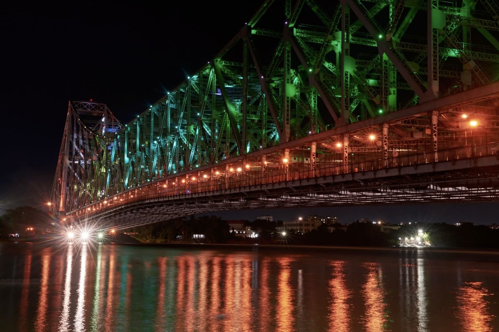 A large illuminated bridge at night, with green and orange lights reflecting on the calm water below, invites you on a holiday adventure. This vibrant metal structure spans across a river, with distant city lights twinkling like a magical LGBTQ+ Christmas destination in the background. Renowned for being one of the best gay Christmas destinations, this enchanting scene promises an unforgettable festive experience filled with inclusivity and celebration.