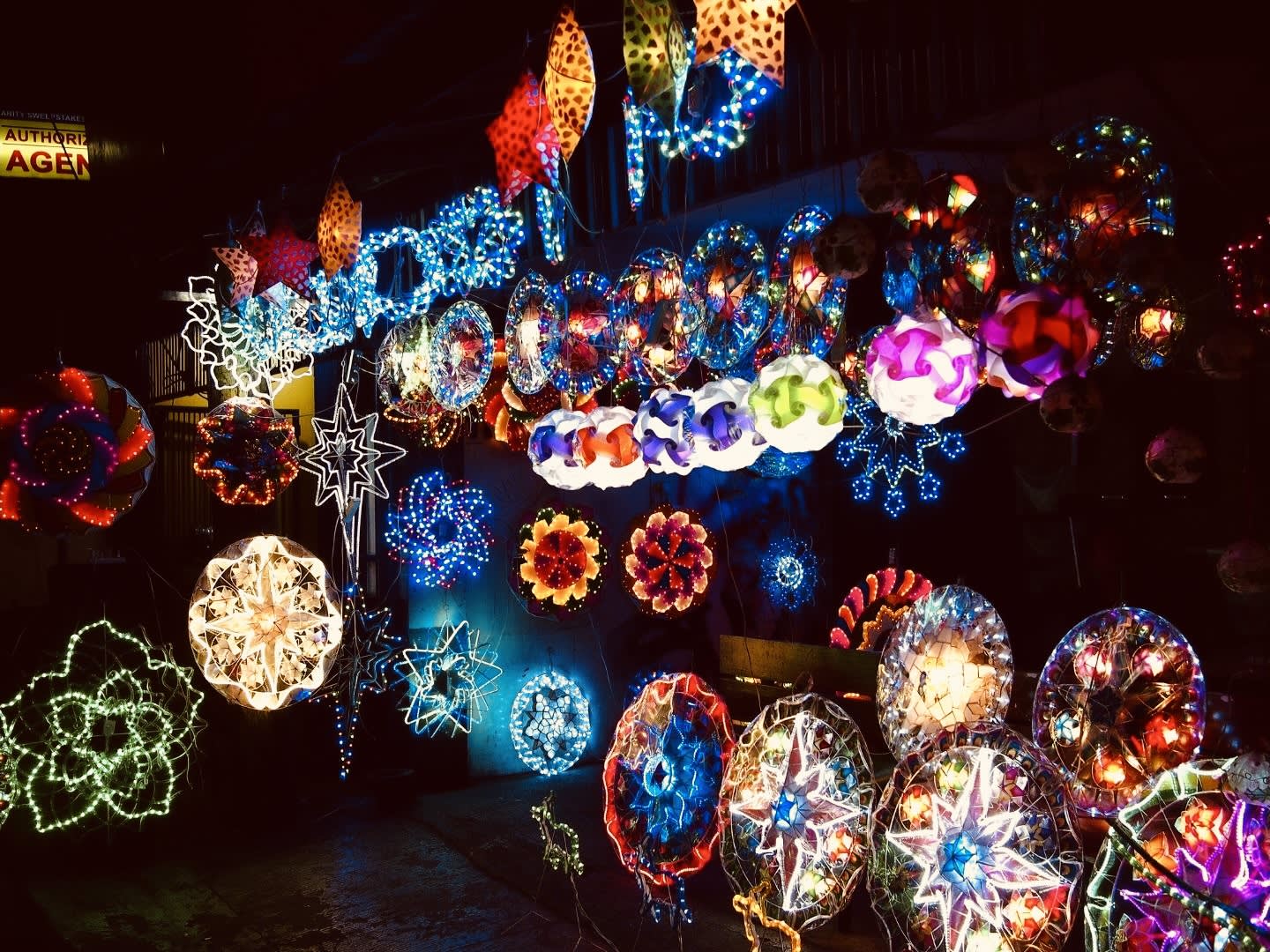 A vibrant display of variously shaped, colorful illuminated lanterns and lights hangs in the dark, capturing the festive spirit and creating a lively atmosphere. The lights form stars and circular patterns in a range of bright colors, reminiscent of the enchanting decorations found at popular gay Christmas destinations around the world. These destinations are celebrated for their welcoming communities and spectacular holiday celebrations that embrace diversity and inclusivity during this joyous season.